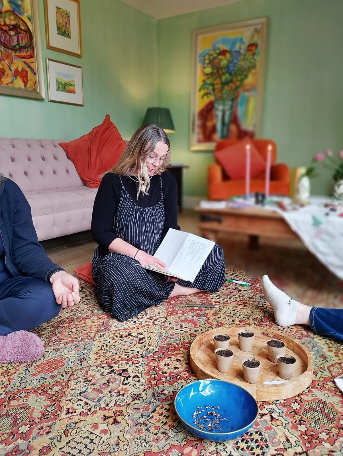 A woman with glasses and blonde hair, sitting cross-legged on a patterned rug, reading a book inside a cozy, colorful living room. There are several art pieces on the green walls, a pink tufted sofa with red pillows, an orange armchair, and a large framed painting of a still life on the wall. A wooden table with some candles and a tablecloth is in the background, and a person’s legs with socks are visible in the foreground.
