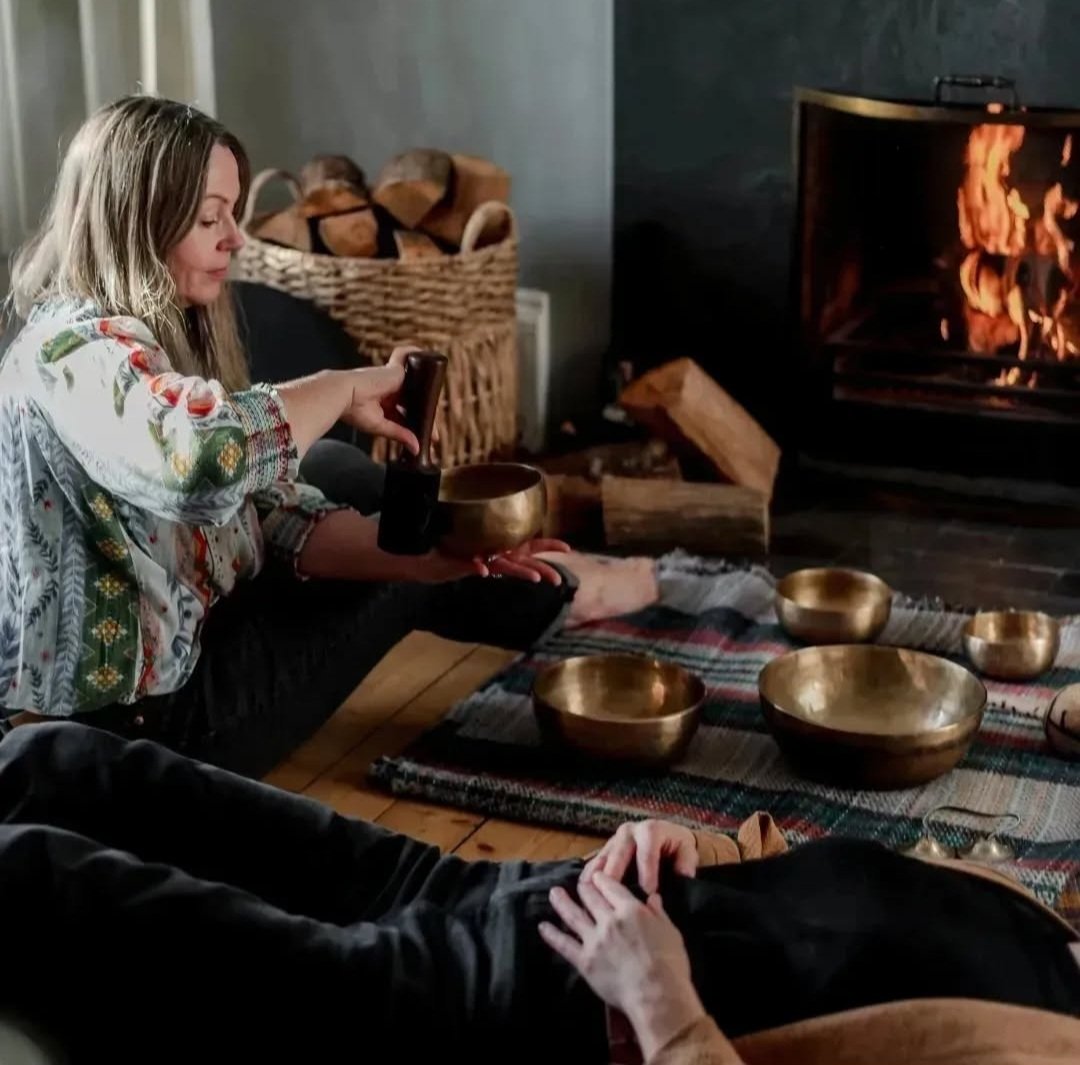 A woman sitting on the floor near a fireplace, taking a photo with her phone, with metal singing bowls on a table, fire burning in the fireplace, and a basket of firewood behind her.