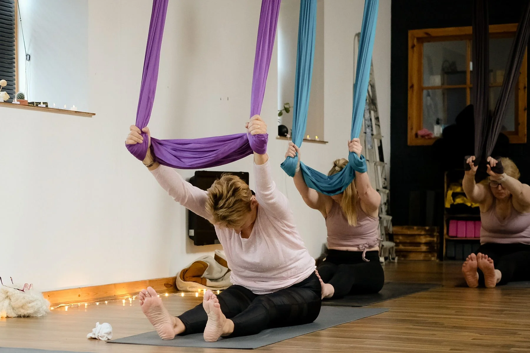 Three women participate in an aerial yoga class, practicing poses with colorful silk hammocks, with two women in the foreground sitting on the floor and the woman on the left holding a purple hammock overhead.