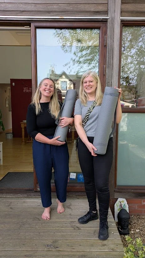 Two smiling women standing outside a building, each holding a rolled yoga mat. One woman is barefoot and dressed in a black top and blue pants, while the other woman is wearing black pants, black shoes, and a gray shirt.