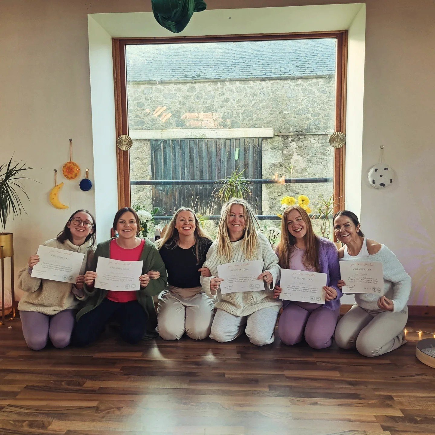 Six women sitting on the floor in front of a large window, holding certificates and smiling.
