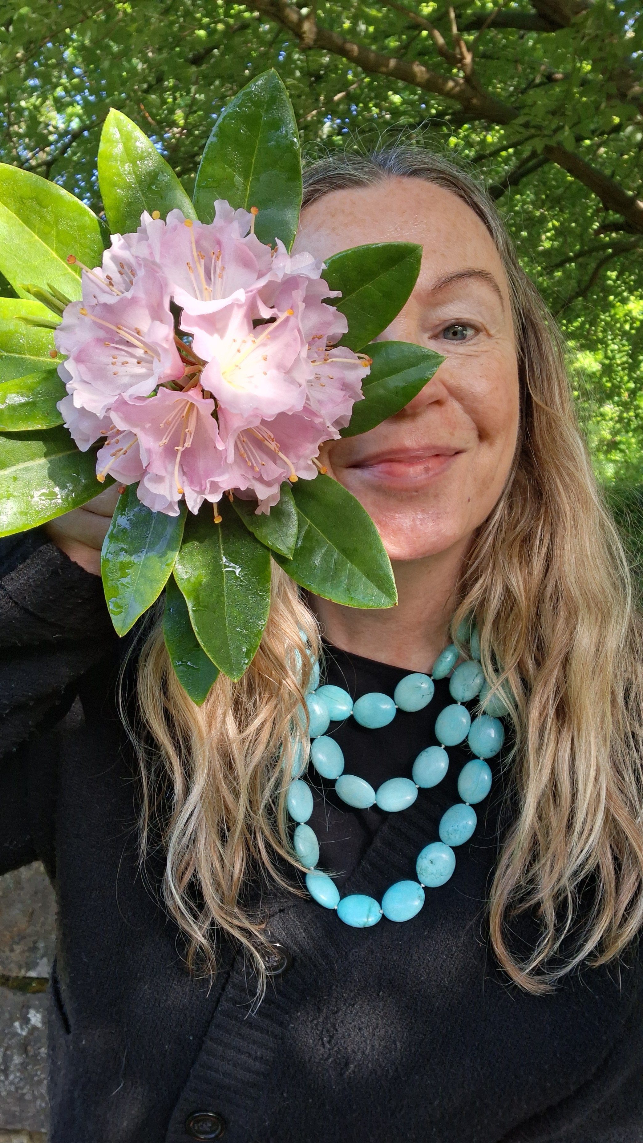 A woman holding a pink rhododendron flower with green leaves, smiling. She has long blonde hair, blue eyes, and is wearing a black jacket and a turquoise beaded necklace.