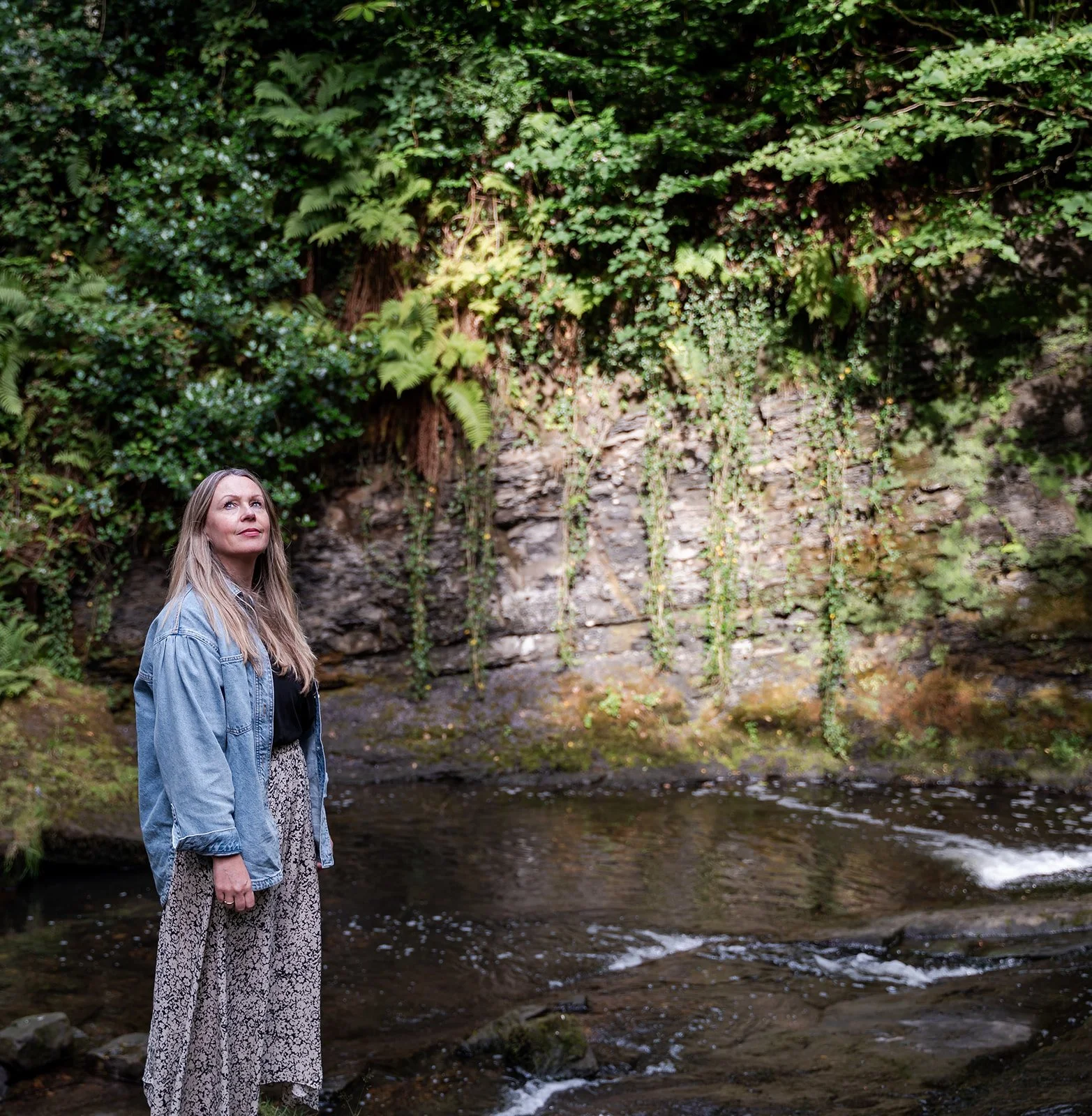 A woman with long blonde hair wearing a denim jacket and patterned dress standing by a creek with rocks and flowing water, with a background of green foliage and trees.