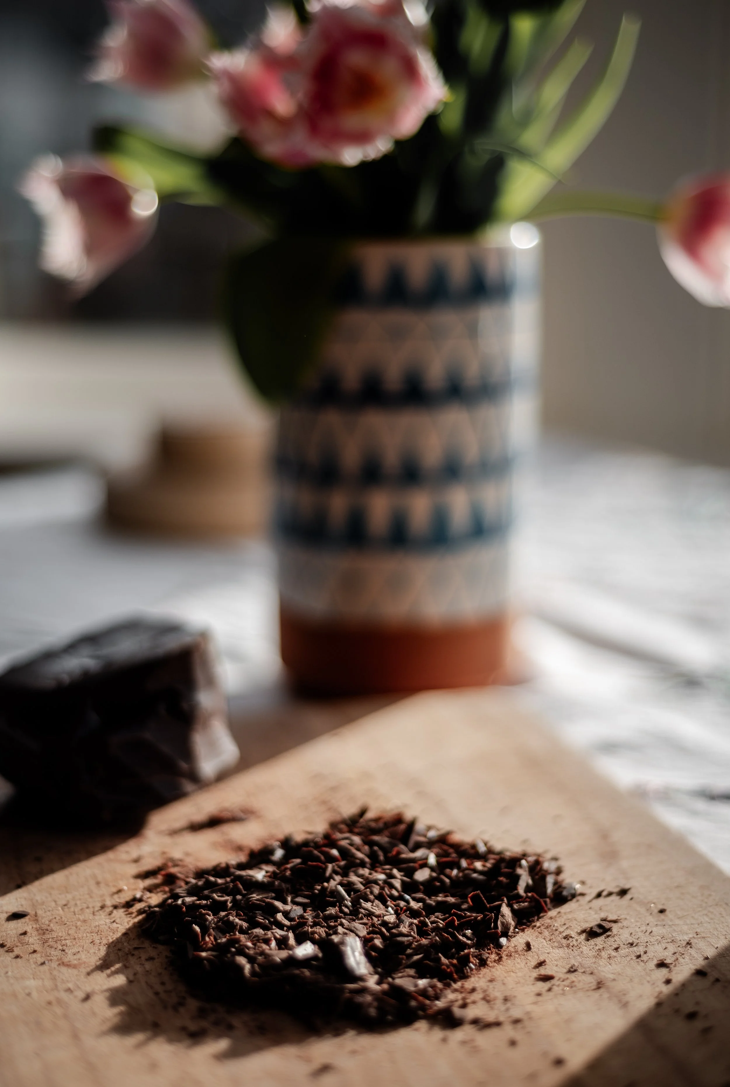 Close-up of dark chocolate shavings and cocoa nibs on a wooden surface, with a blurred background including a chocolate bar and a patterned planter with pink flowers.