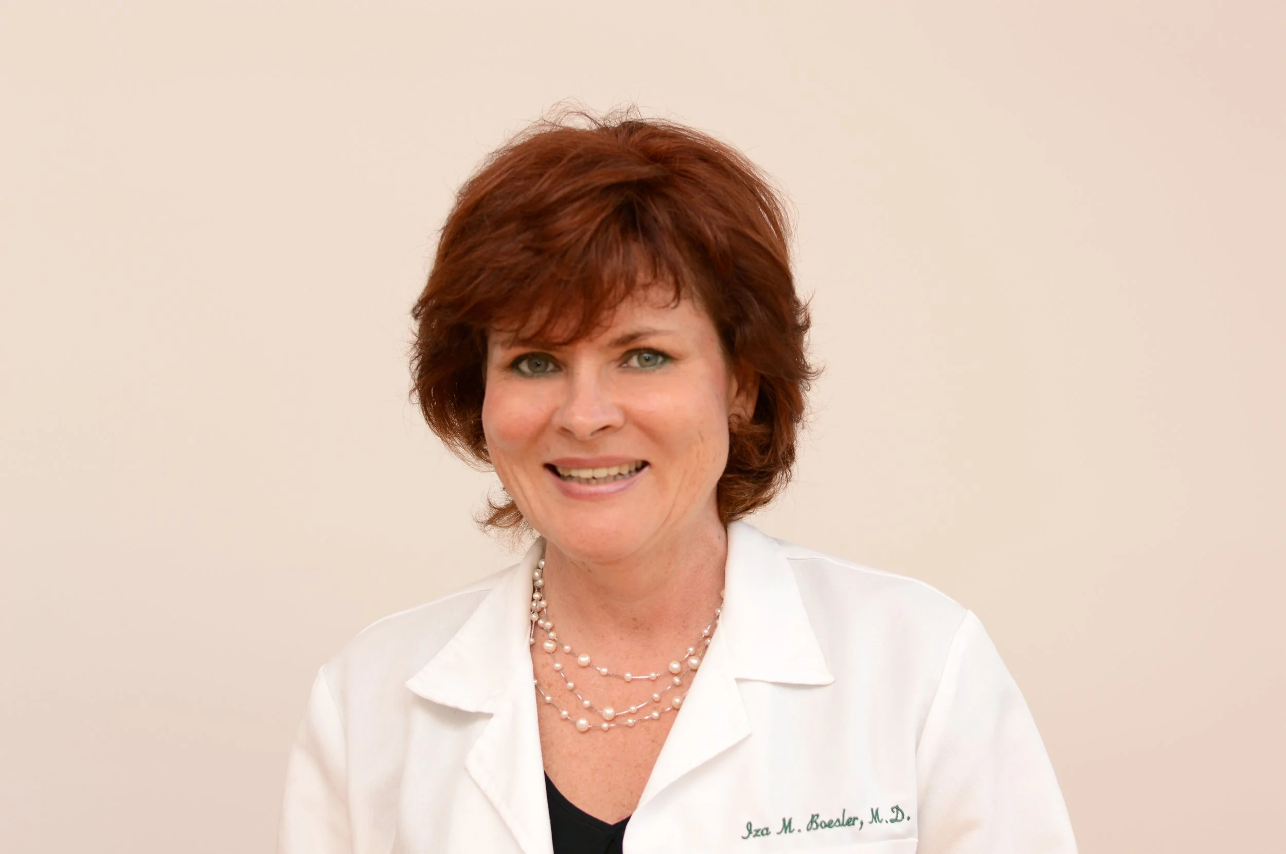 A smiling woman with short red hair wearing a white coat and a pearl necklace, against a light beige background.