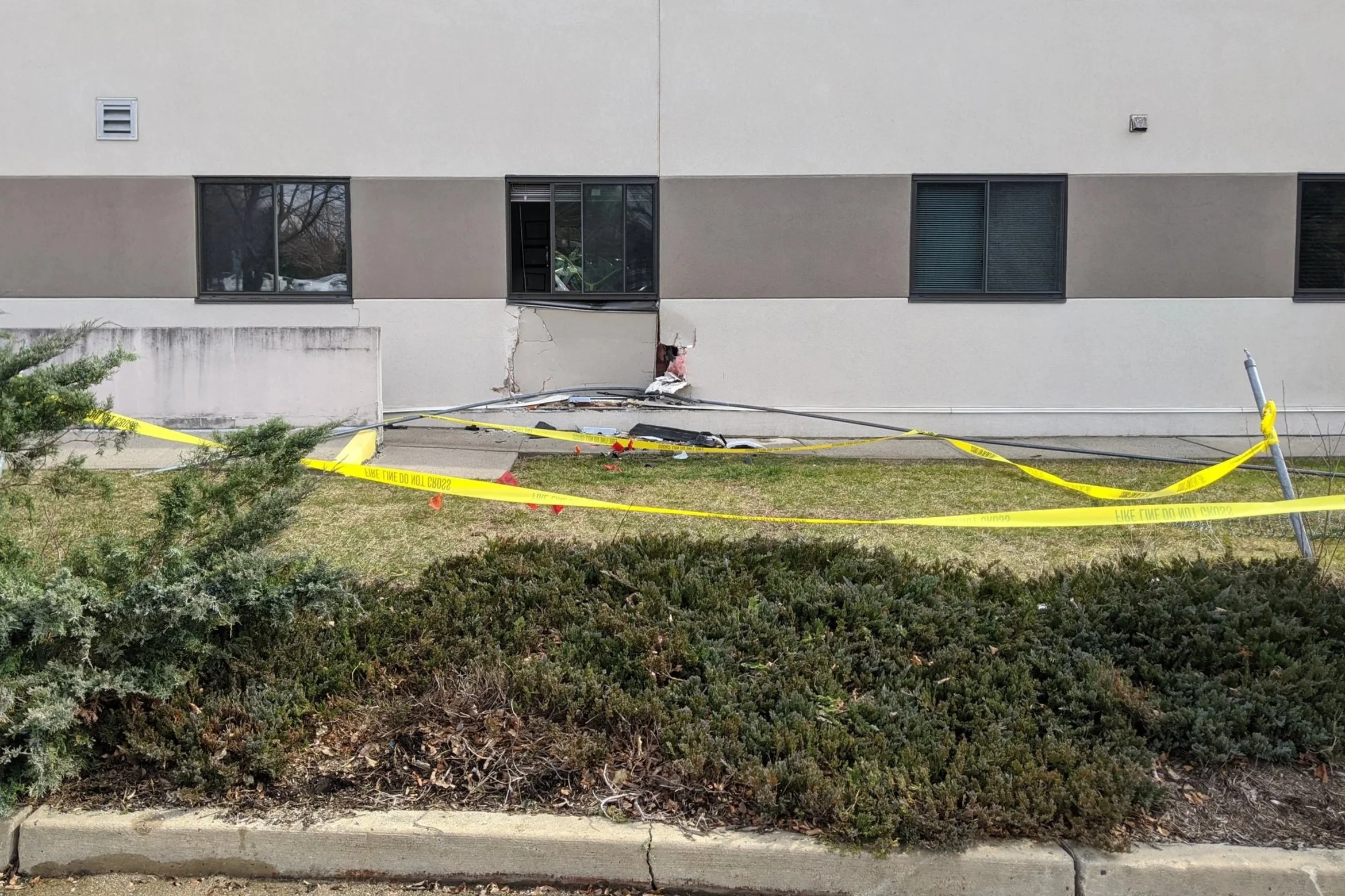 Exterior of a building with damaged wall and broken window, yellow caution tape, and construction debris on the ground.