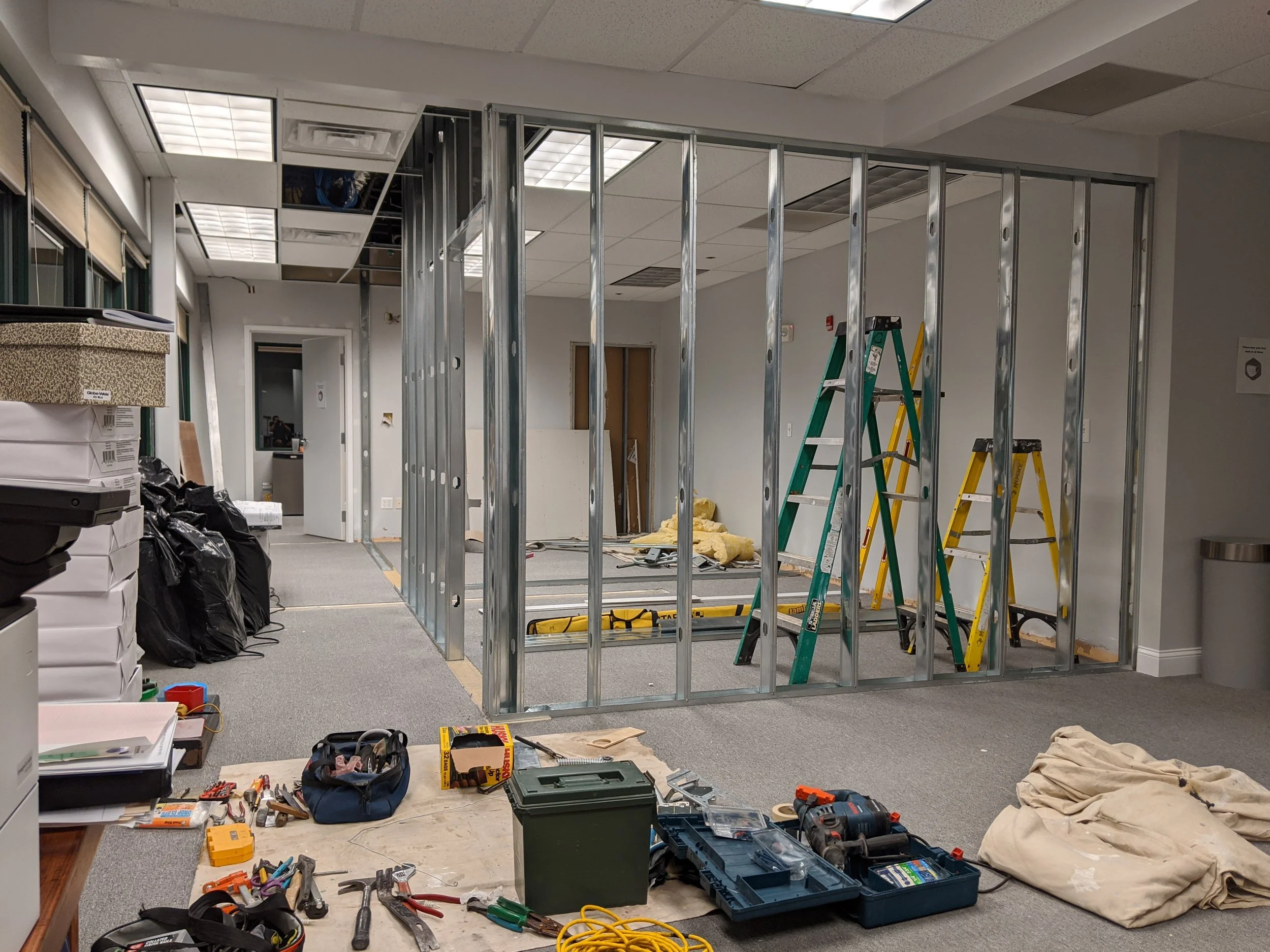 An office in the process of renovation, with metal wall framing installed, ladders, construction tools, supplies, and materials scattered around.