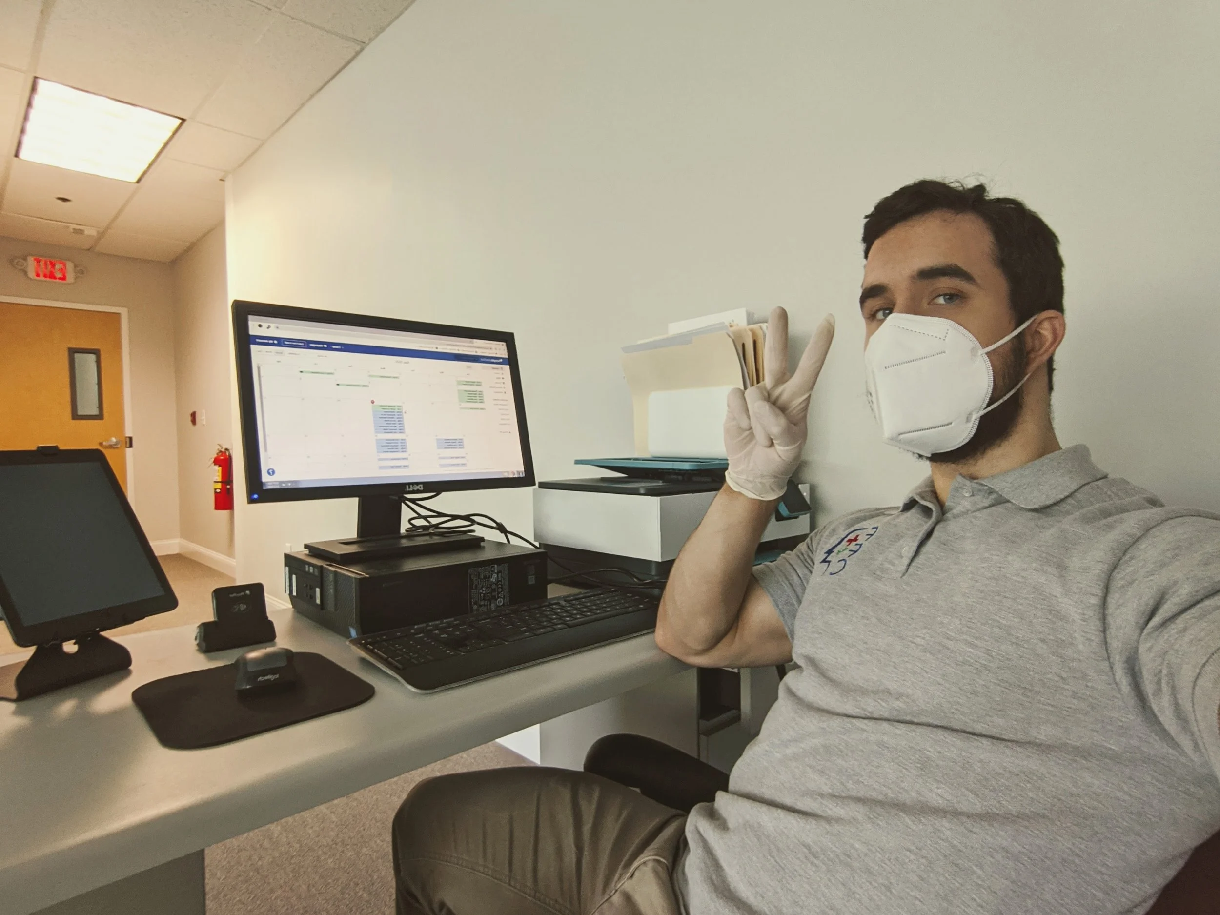 A man wearing a white face mask and gray polo shirt making a peace sign at a desk with computer monitors, located in an office with a closed door, ceiling lights, and peach-colored walls.