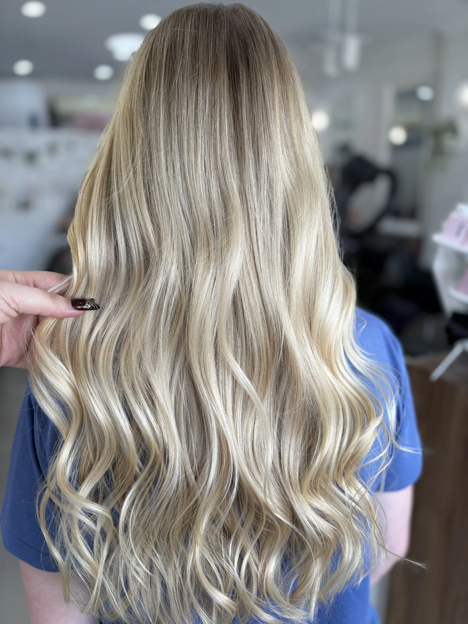 A person with long, wavy blonde hair wearing a blue shirt, seen from behind in a hair salon.