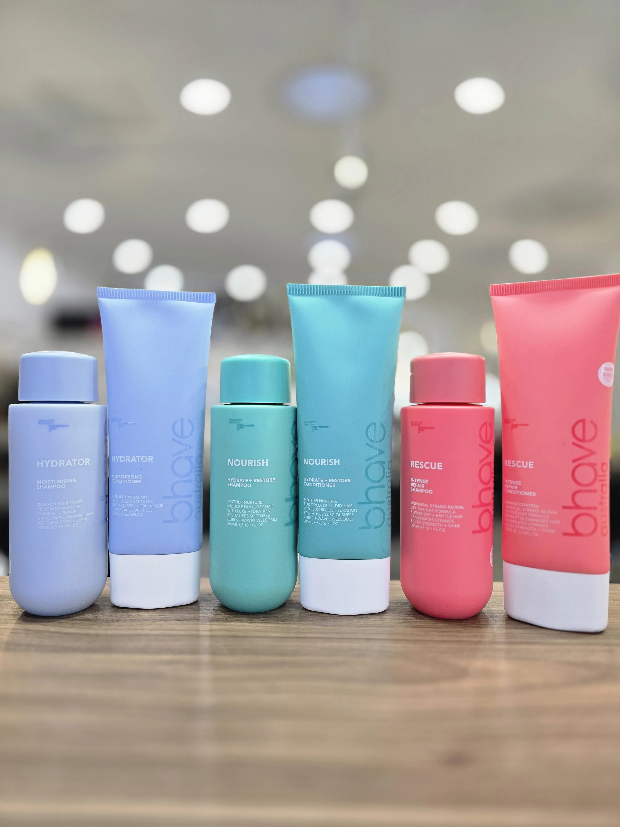 A row of colourful Bhave hair care bottles in blue, green, and pink on a wooden surface, with a blurred store ceiling and lights in the background.