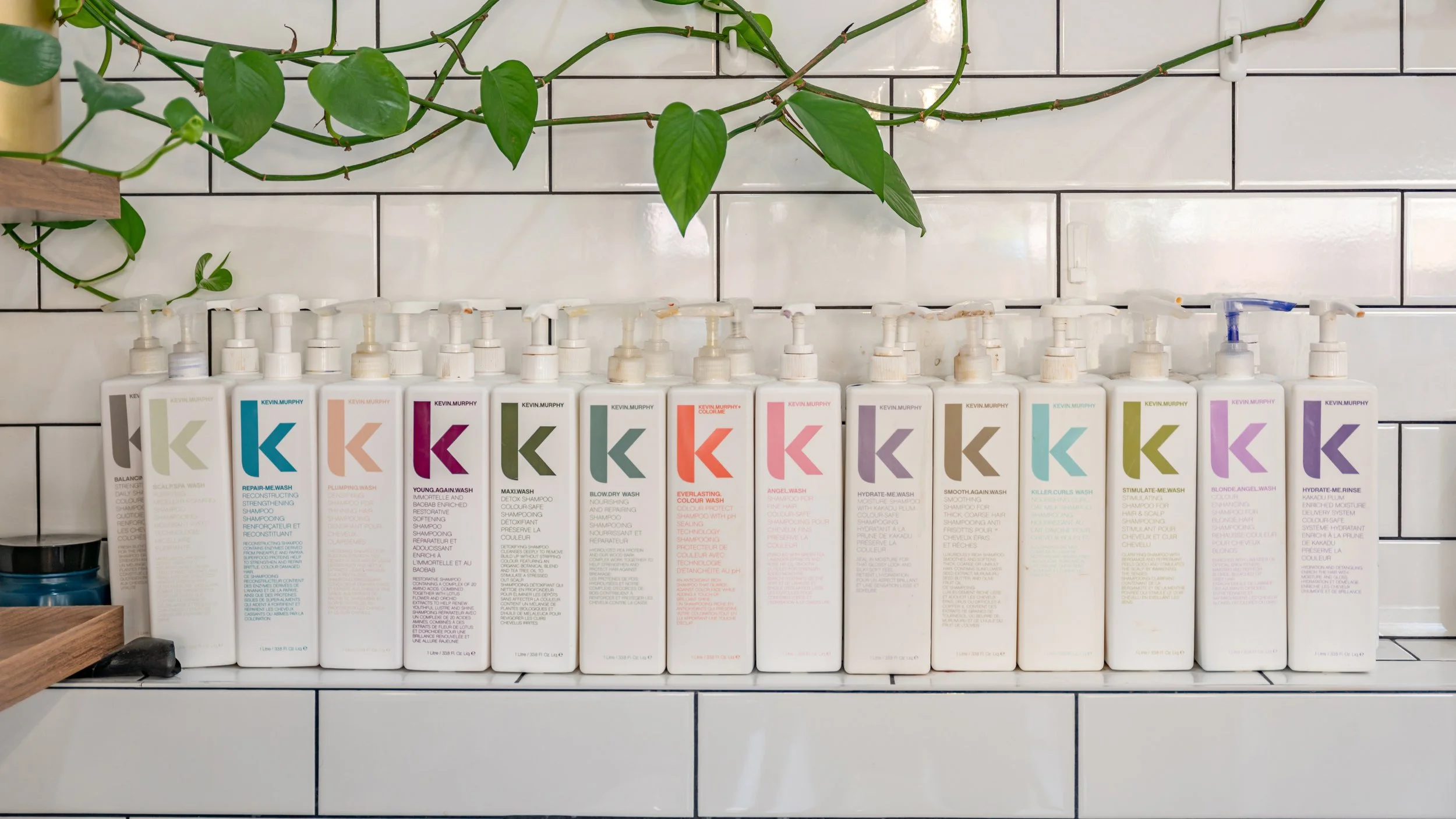 Collection of Kevin Murphy hair care product bottles arranged on a shelf against a white tiled wall with some green plant trailing above.