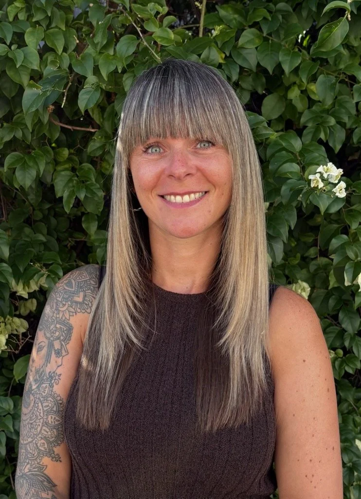 A woman with straight blonde hair and bangs, smiling, standing outdoors in front of green leafy bushes with white flowers, wearing a sleeveless dark top.