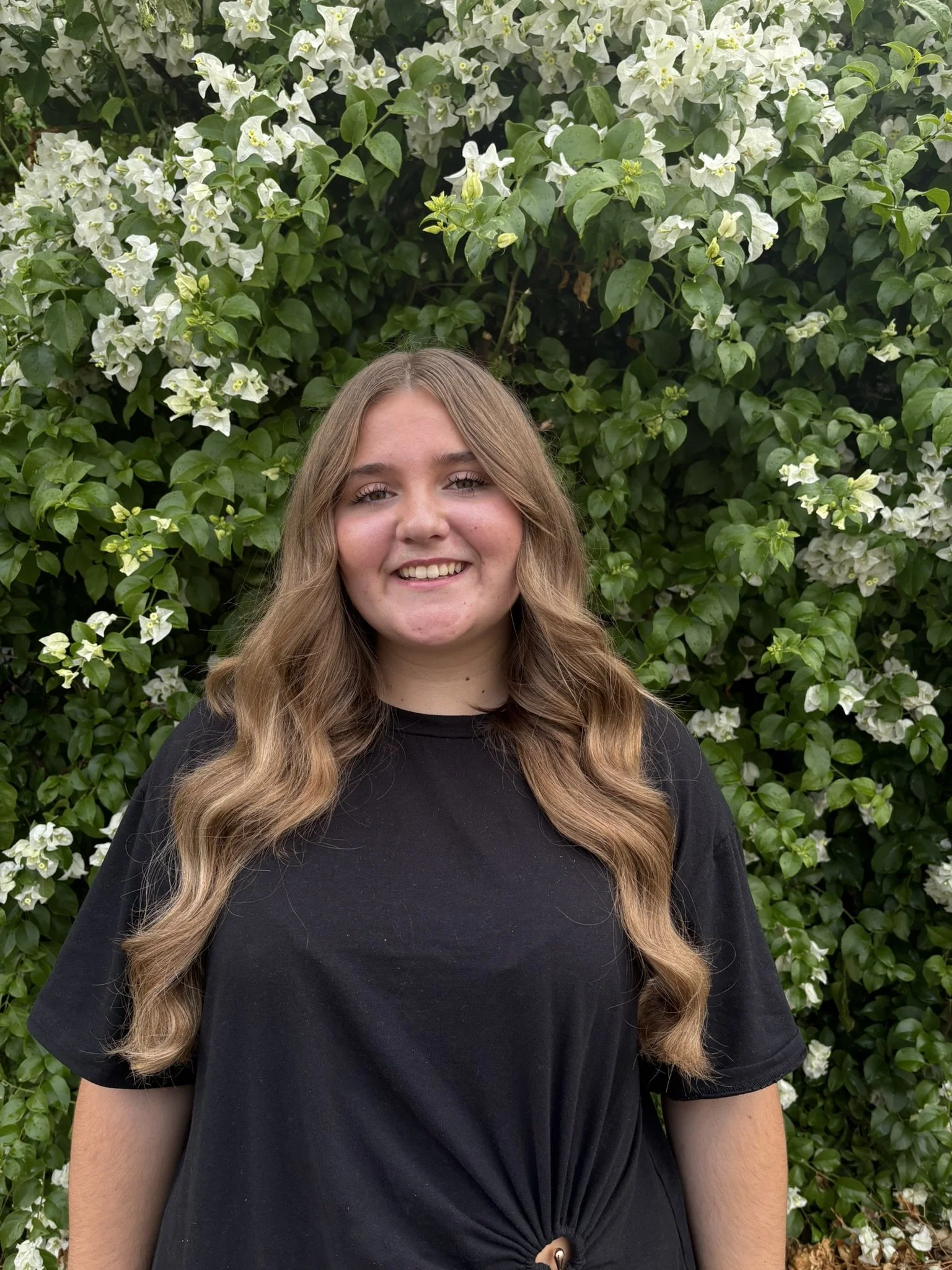 A young woman with long wavy hair smiling, wearing a black t-shirt, standing in front of a lush green bush with white flowers.
