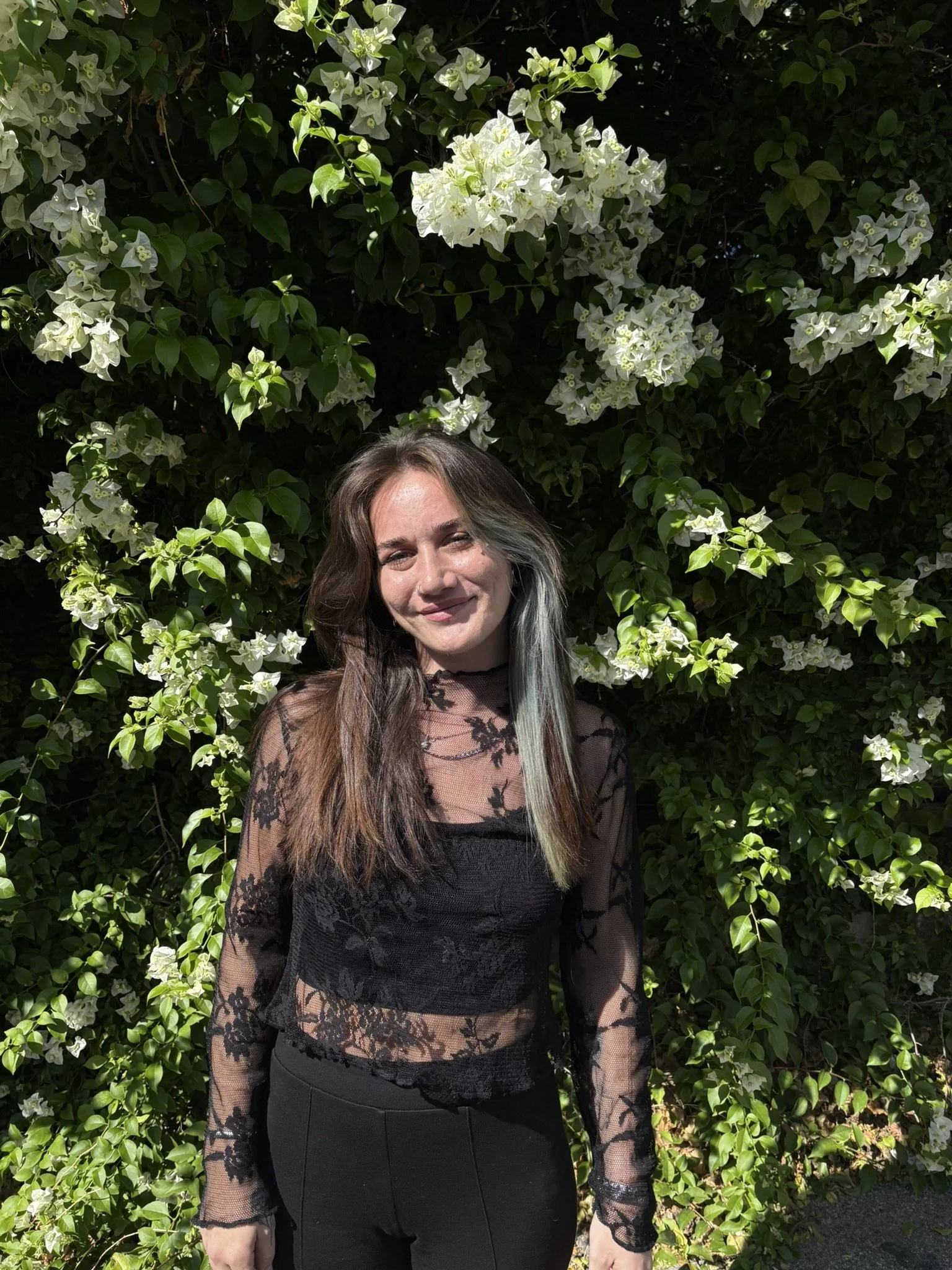 A woman standing in front of lush green foliage with white flowers. She has long brown hair with dyed portions, and is wearing a black lace top and pants. Sunlight is shining on her face.