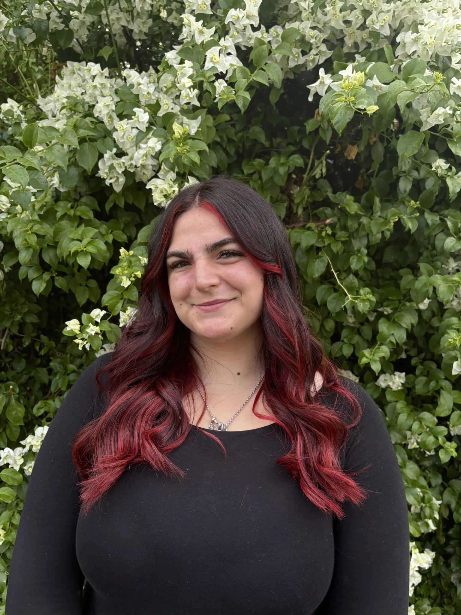 A young woman with long, dark hair with red highlights, smiling, standing in front of a lush green bush with white flowers.
