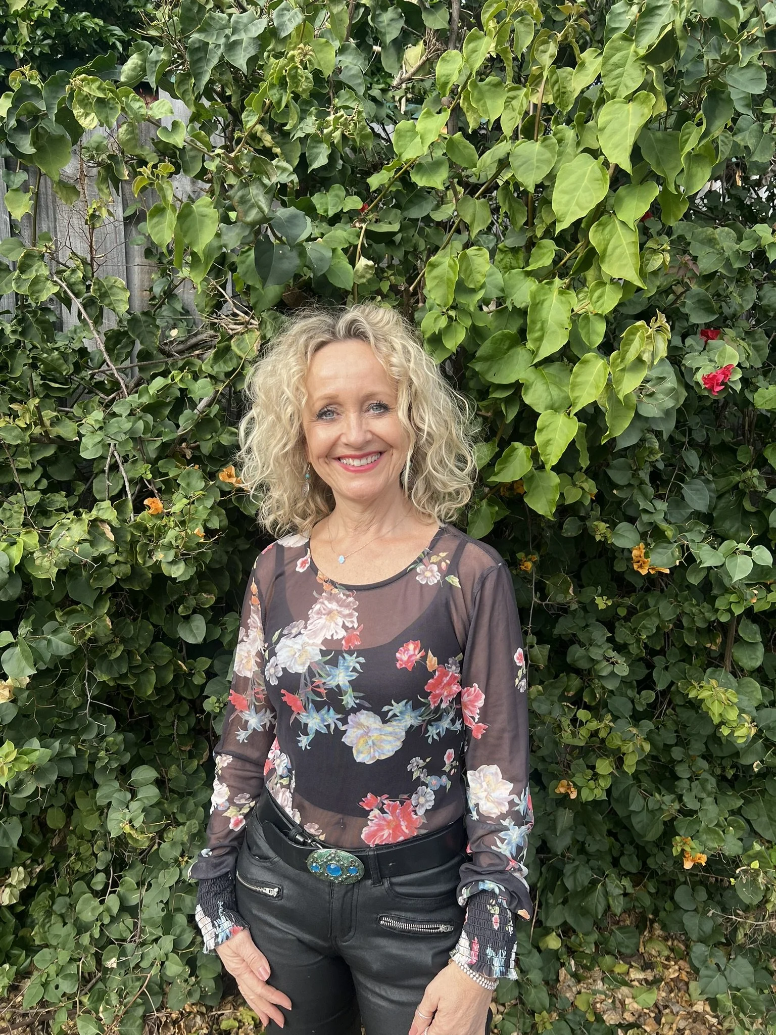 A woman with curly blond hair, wearing a sheer black floral top and black pants, smiling while standing outdoors in front of a lush green leafy hedge with a few pink flowers.