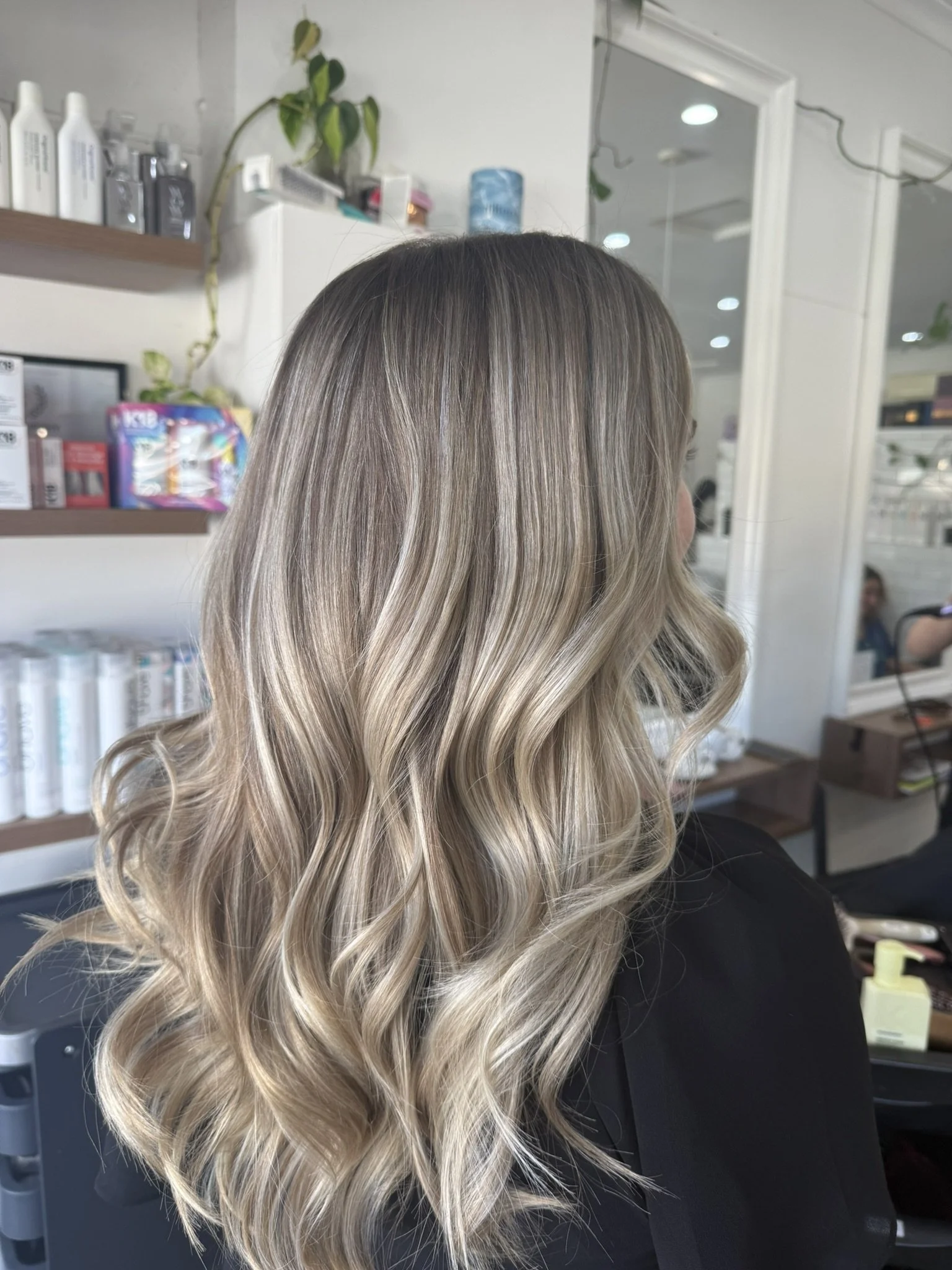 The image shows the back of a woman with long, wavy, blonde hair in a salon. The background includes salon products and a mirror.