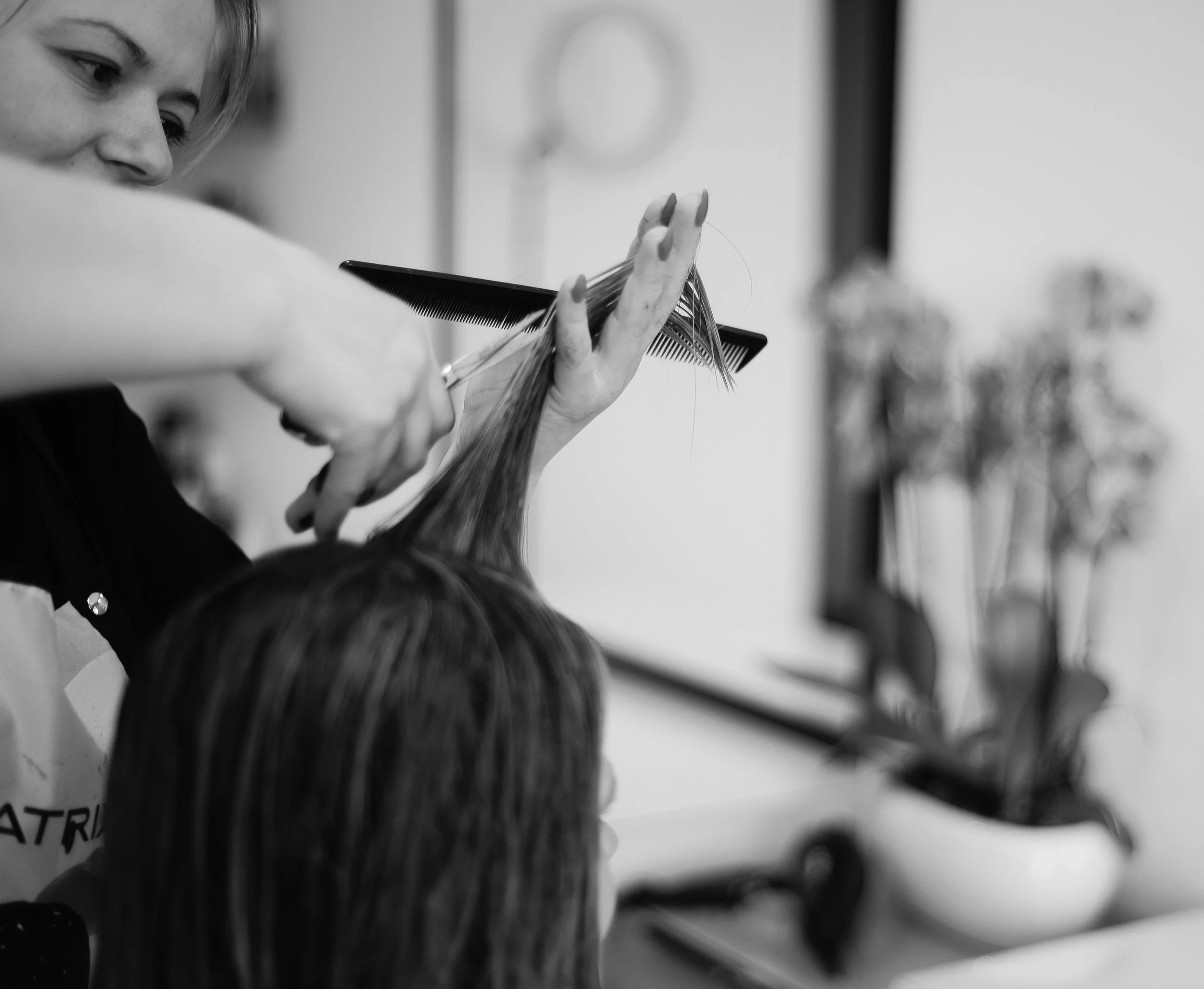 A woman with blonde hair and a focused expression is using scissors and a comb to cut or style a person's hair in a salon setting.