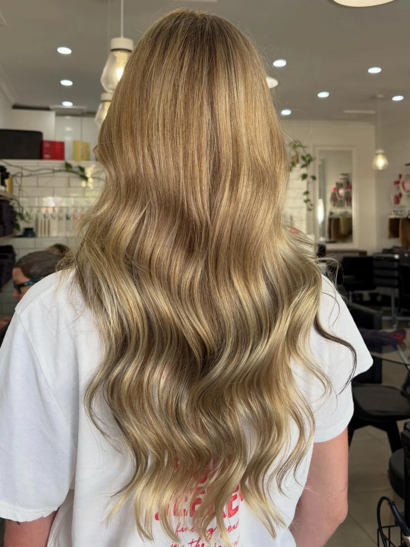 A person with long, wavy blonde hair standing in a salon or store with modern lighting, shelves, and decor in the background.