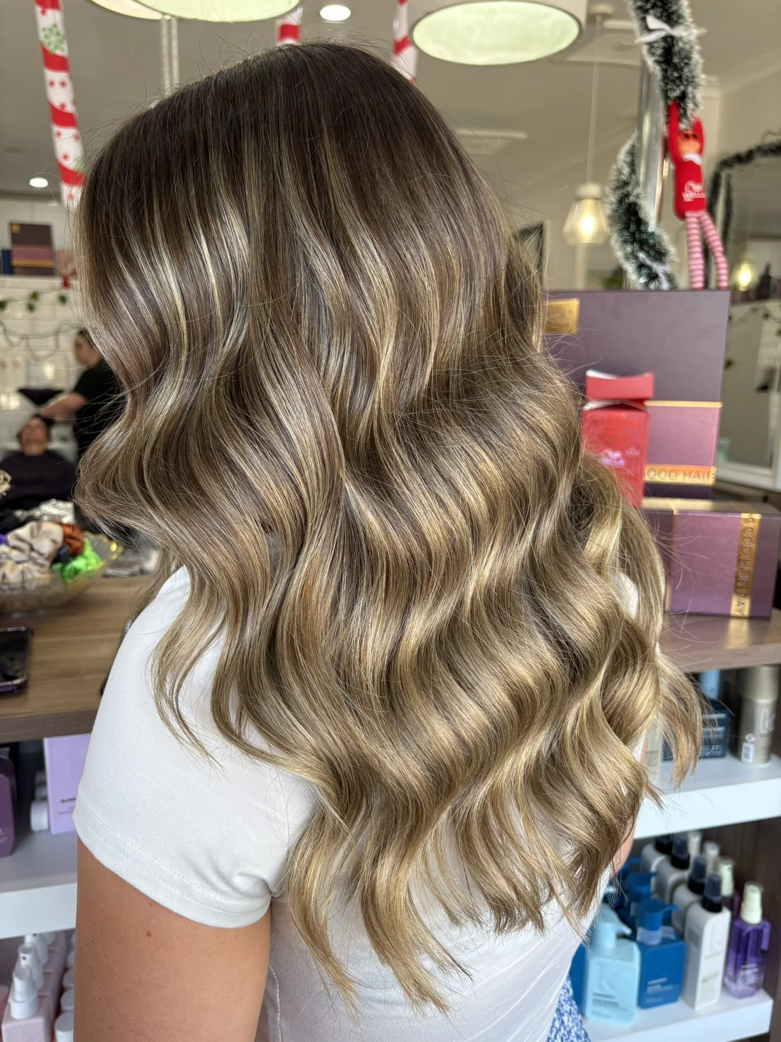 Woman with long, wavy hair featuring blonde highlights and darker roots, standing in a salon with shelves of hair products in the background.
