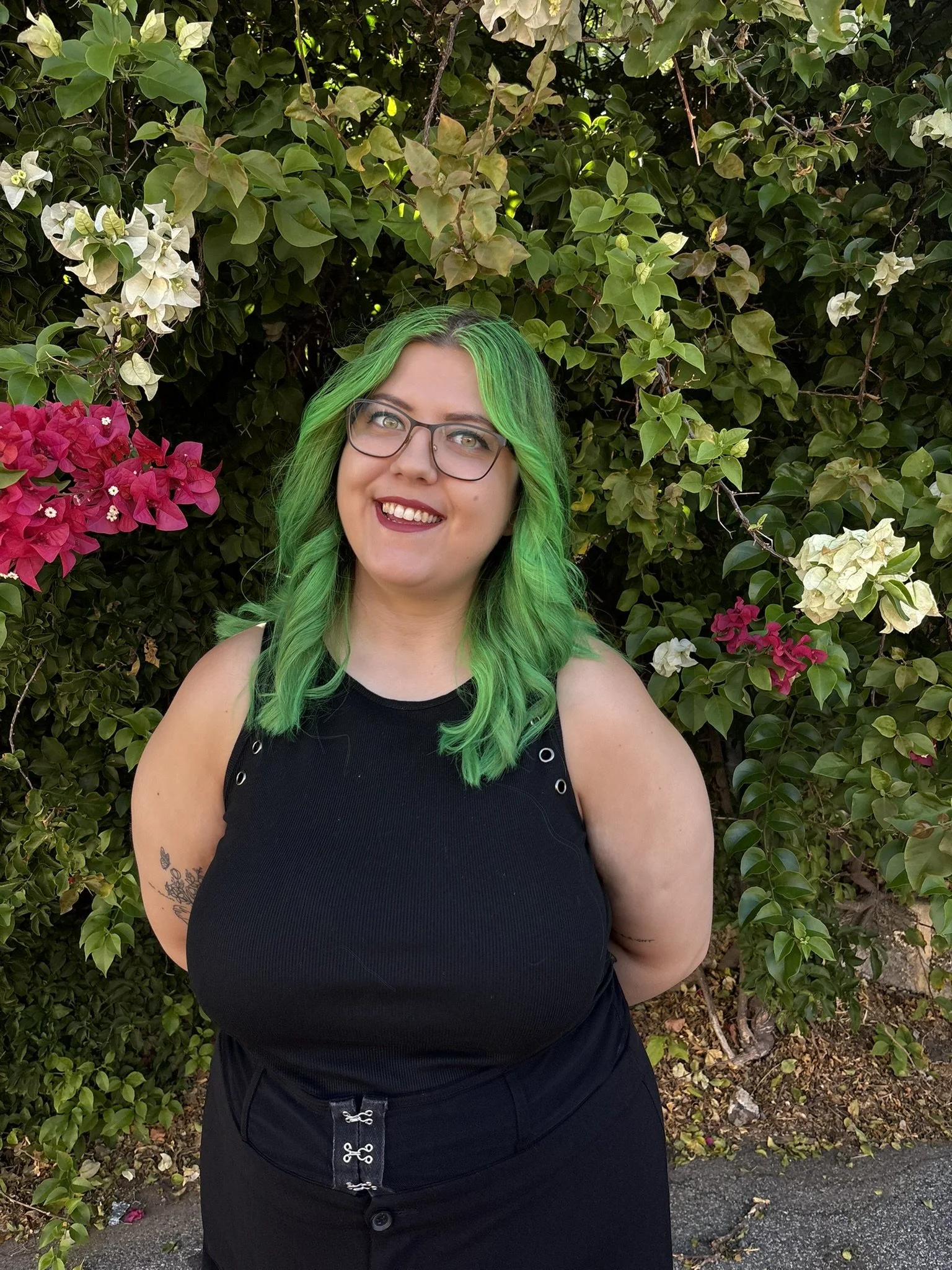 A woman with long green hair wearing glasses and a black top, standing outdoors in front of a bush with pink, white, and green leaves.