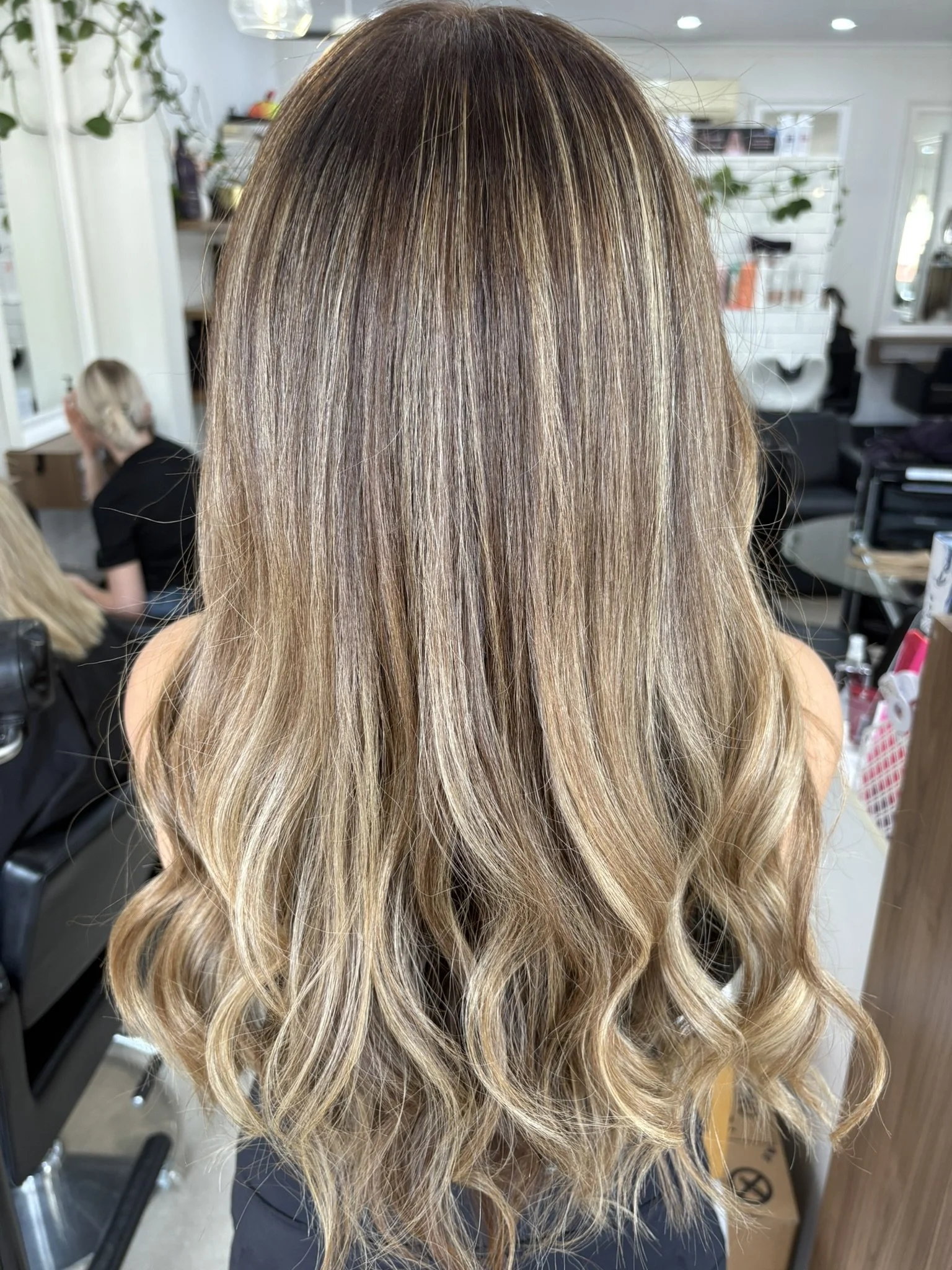Close-up of a woman's head showing newly styled, wavy, highlighted blonde hair with darker roots, in a hair salon.