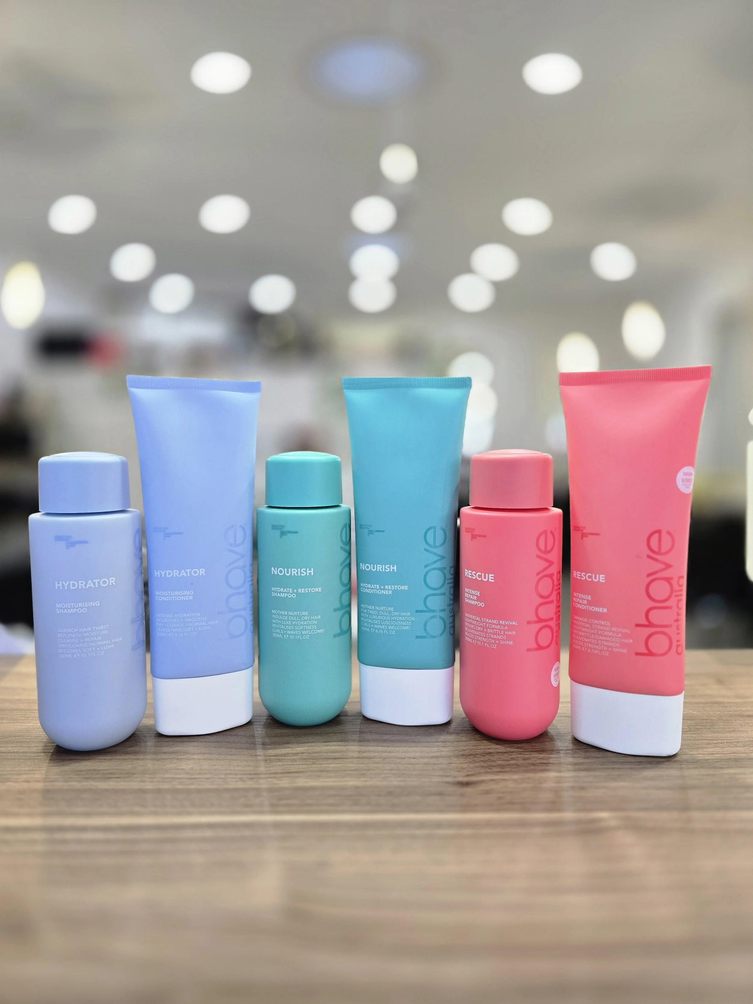 Four Bhave bottles of shampoo and conditioner products in pastel colours on a wooden surface, with a blurred background of ceiling lights.