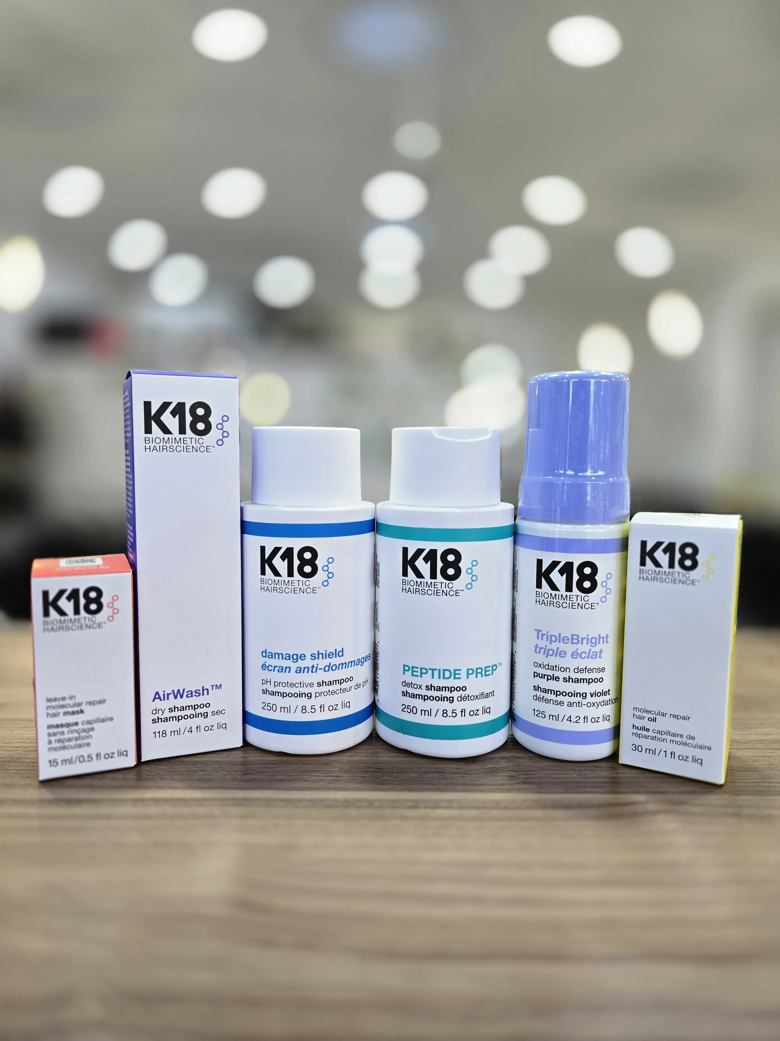 Collection of haircare products including shampoos and a hair oil from the K18 Biometric Hairsience line, placed on a wooden surface with a blurred background.