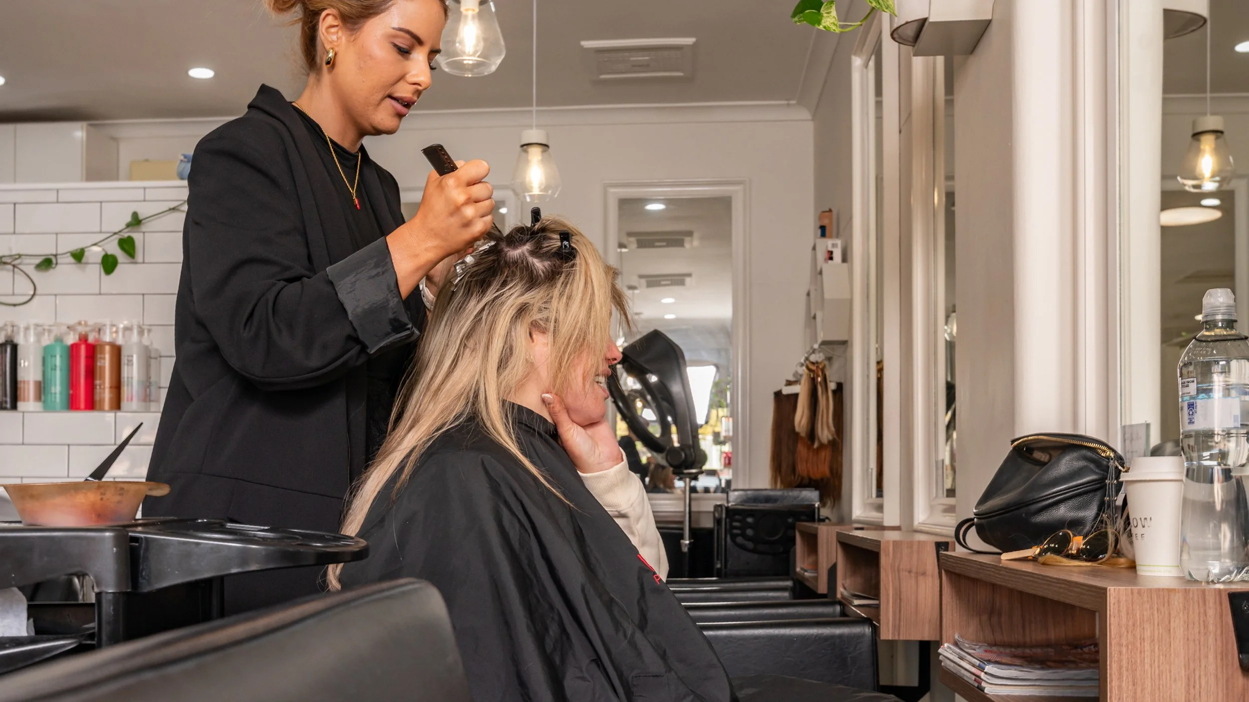 A hairstylist with earrings and a necklace is blow-drying a woman's hair with rollers in a modern salon. The woman is seated facing a mirror, and various hair products and personal items are on the counter.