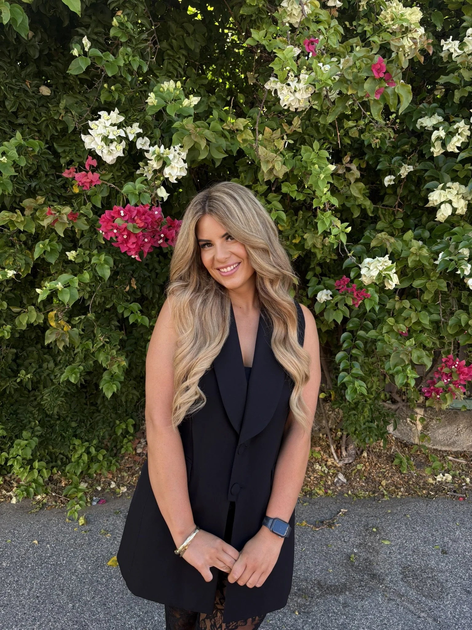 A woman with long, wavy blonde hair smiling in front of a lush green bush with white and pink flowers, wearing a black sleeveless dress and a smartwatch.