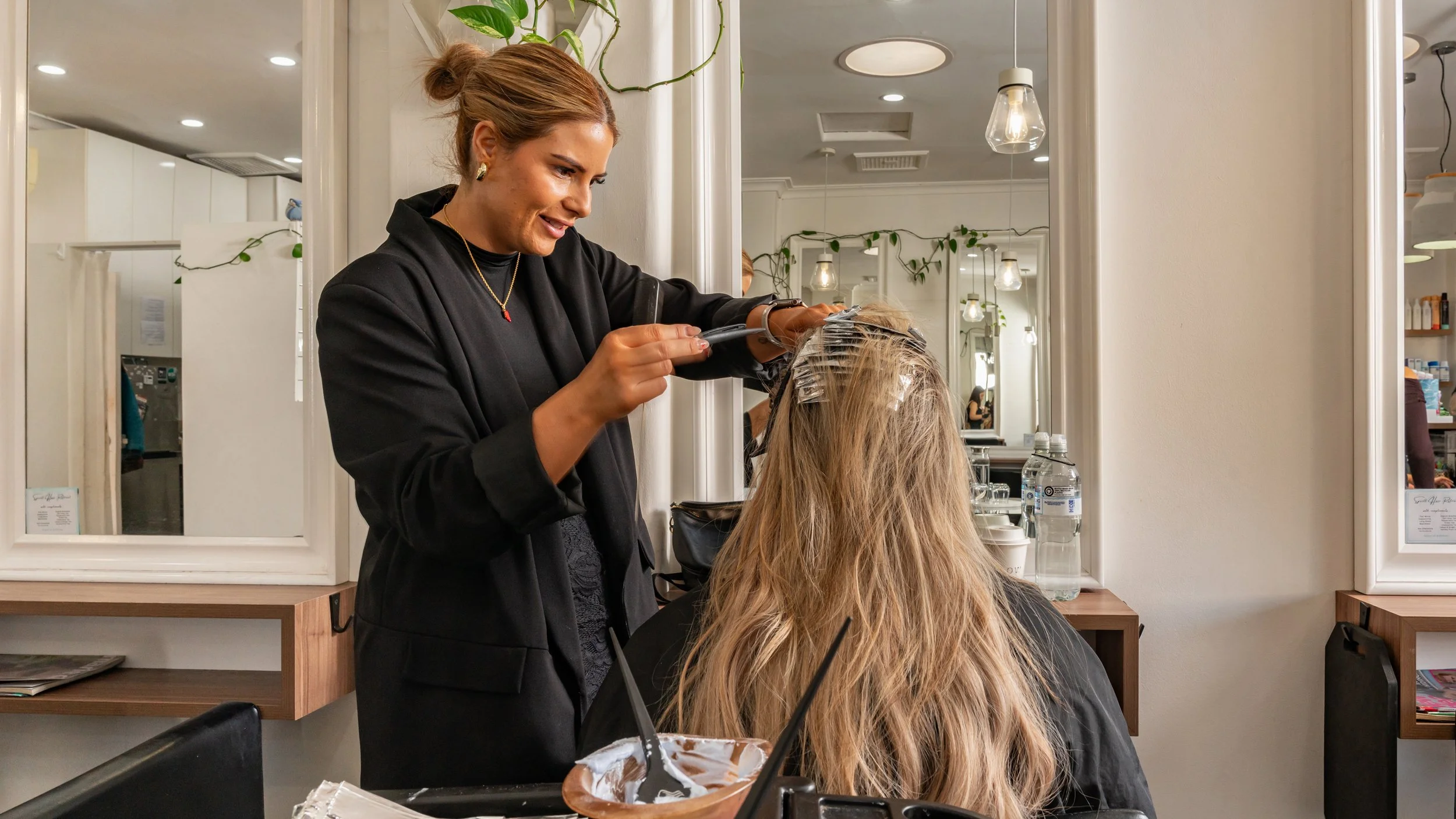 A hairstylist blow-drying a woman's hair at a salon, with mirrors, plants, and salon supplies visible in the background.