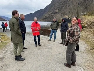 Syv personer står i en gruppe ved en vannkyst under fjell, snakker og smiler.