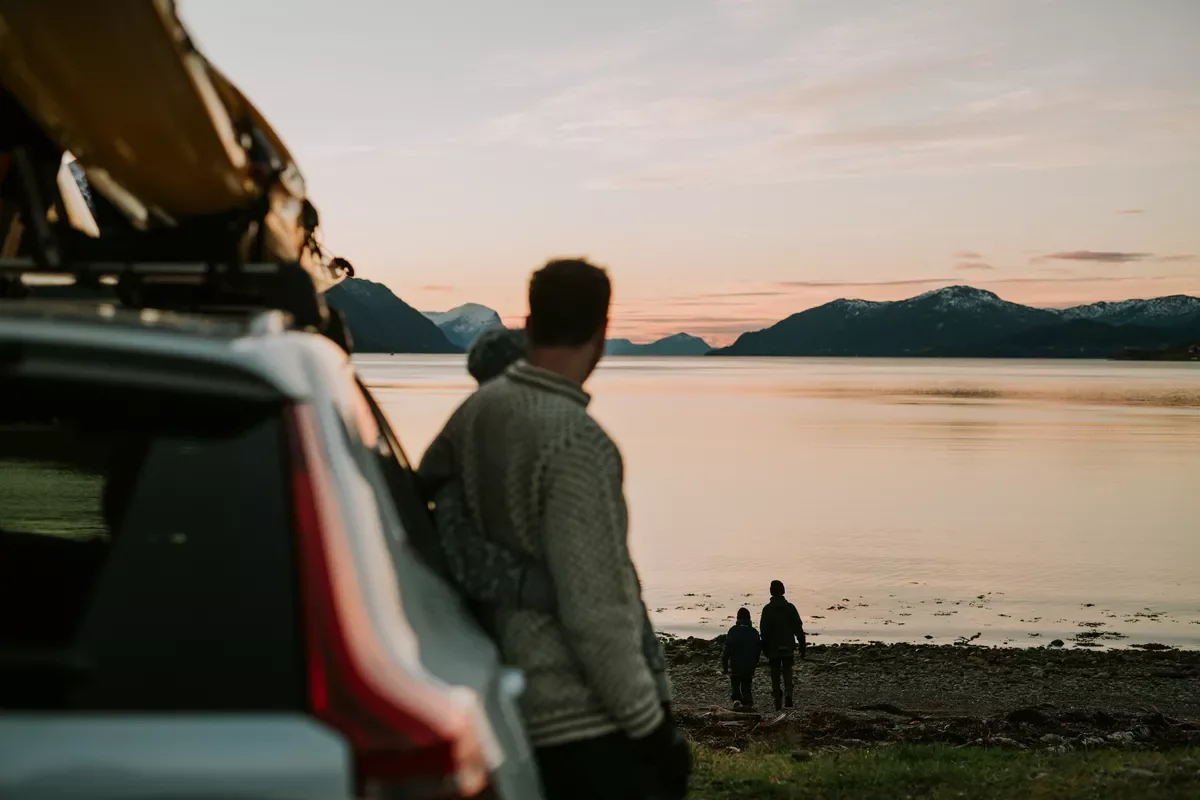 Personer som står ved en innsjø, med fjell i bakgrunnen, under solnedgang, sett fra innsiden av en bil.