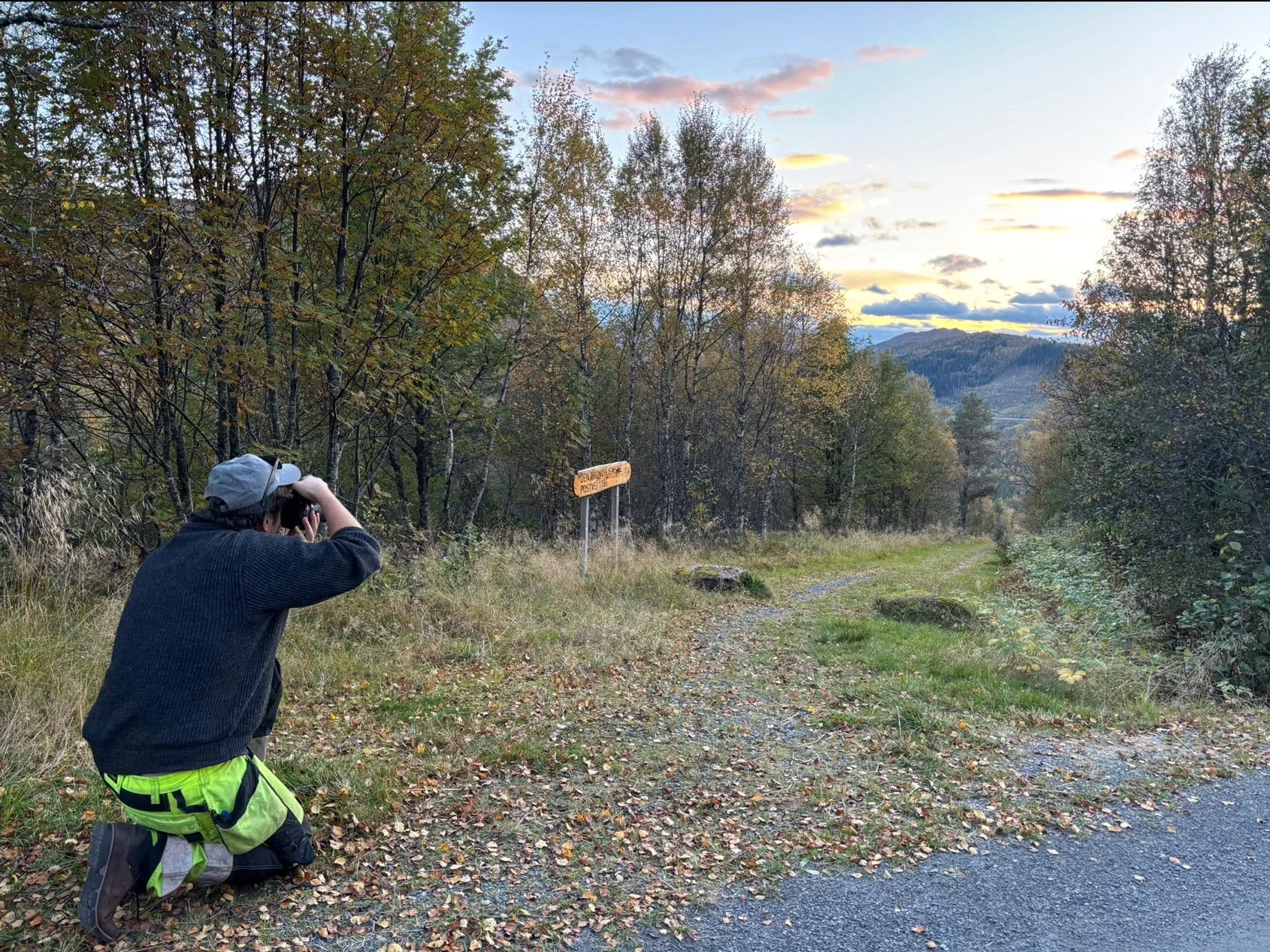 En person med kamerautstyr fotograferer en stien i en skog, rundt høstfargede trær, med fjell og skyer i bakgrunnen, under en delvis skyet himmel.