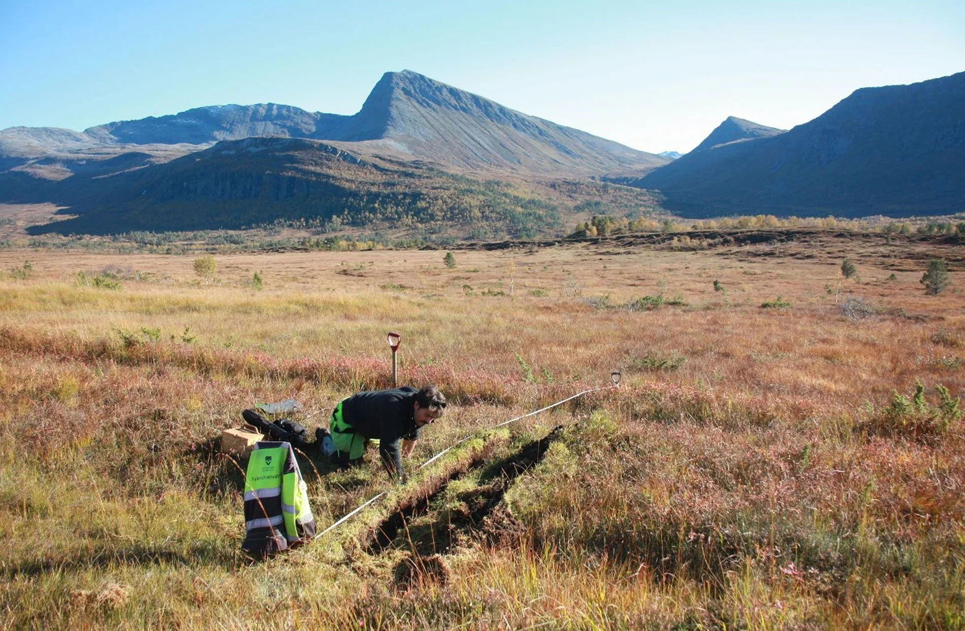 En person bøyer seg ned i en åpen mark med høstfargede planter, omgitt av fjell og klare blå himmel, mens de håndterer noe på bakken.