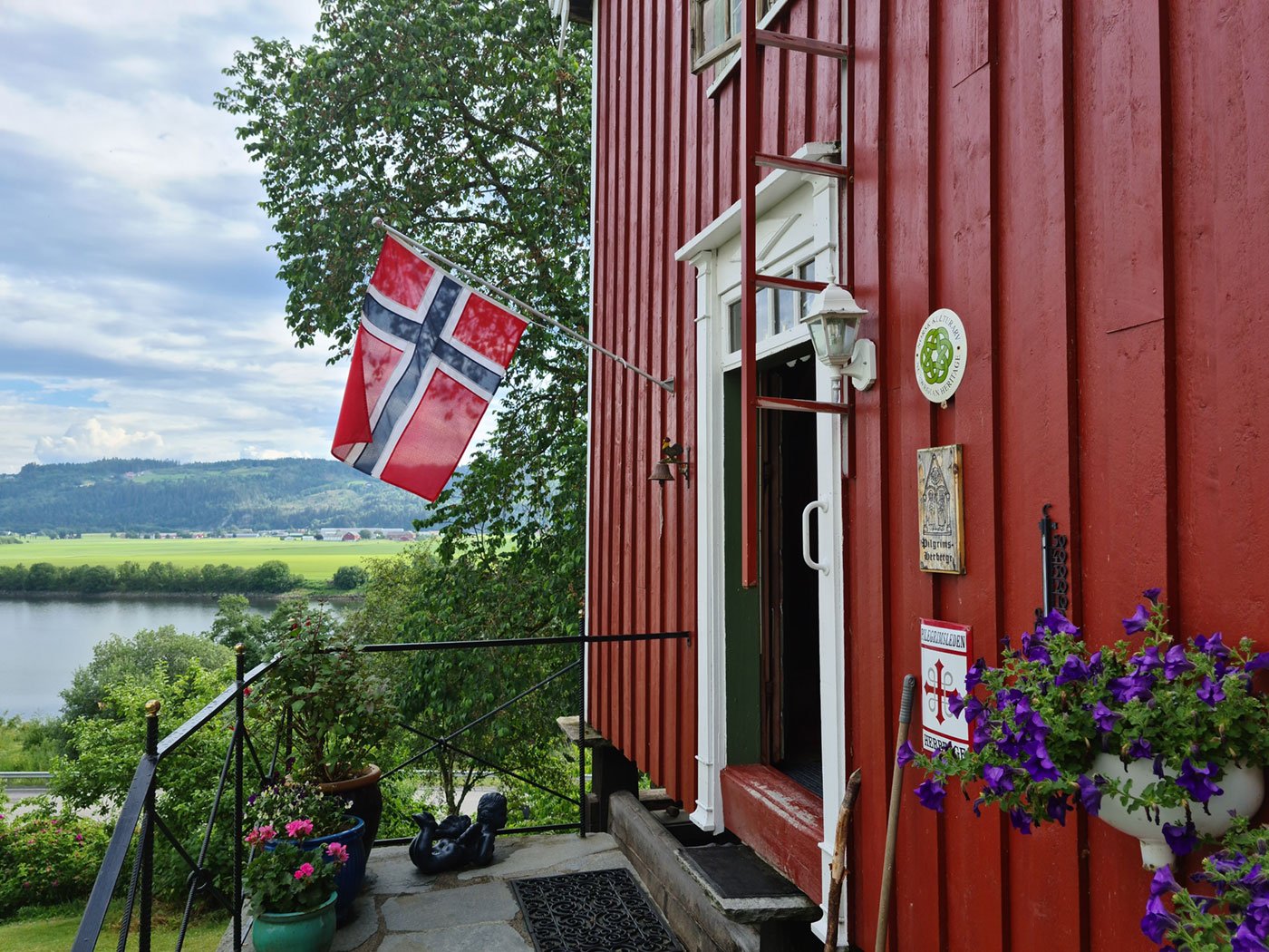 Utsikt fra en terrasse foran en rød panelhus med en norsk Flagget hengt utendørs, blomster i Potte og utsikt over eiendommen og landskapet rundt, med vann og grønne åser i bakgrunnen.