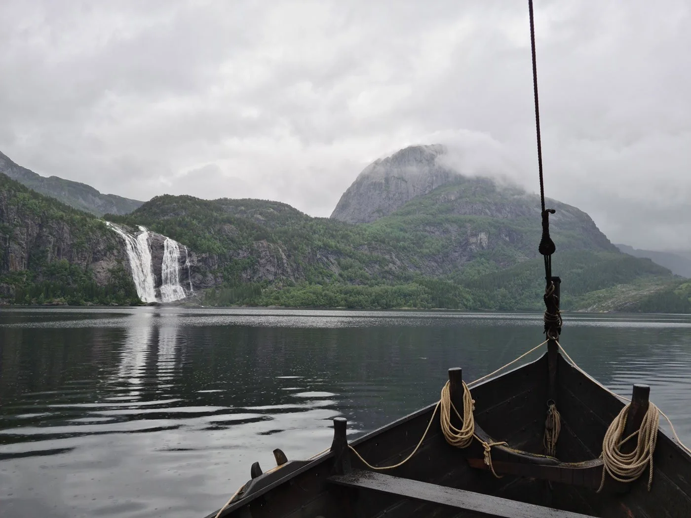 Fyrtårn av en båt på innsjø omgitt av fjell med fossefall, noen skyer og lavt skydekke.