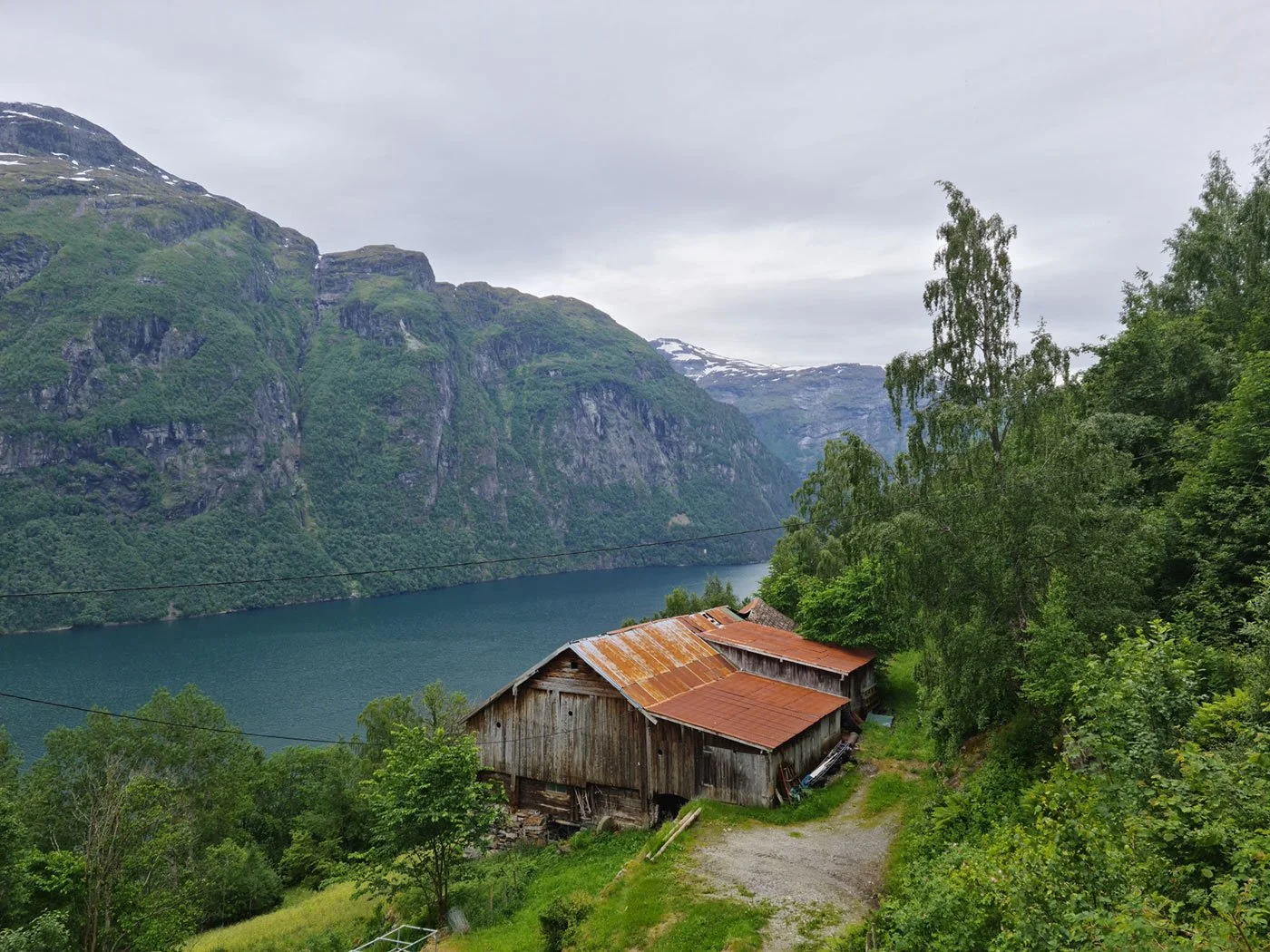 Et gammelt værbit hønsehus ved en innsjø omgitt av grønne trær og fjell med snø på toppen.
