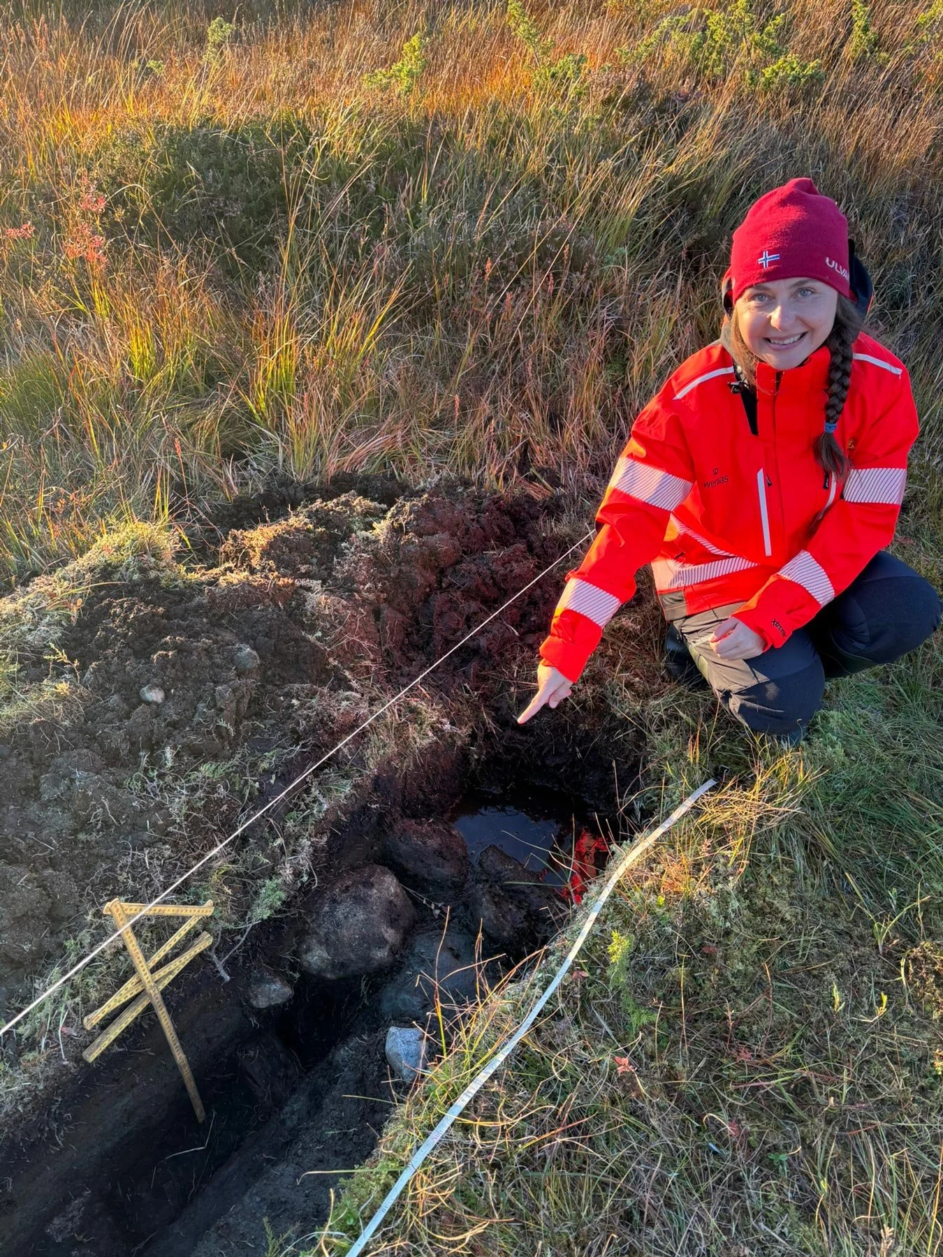 En kvinne i rød fjellutstyr, smilende, knepende ved en jordhule i et gressområde under solnedgang