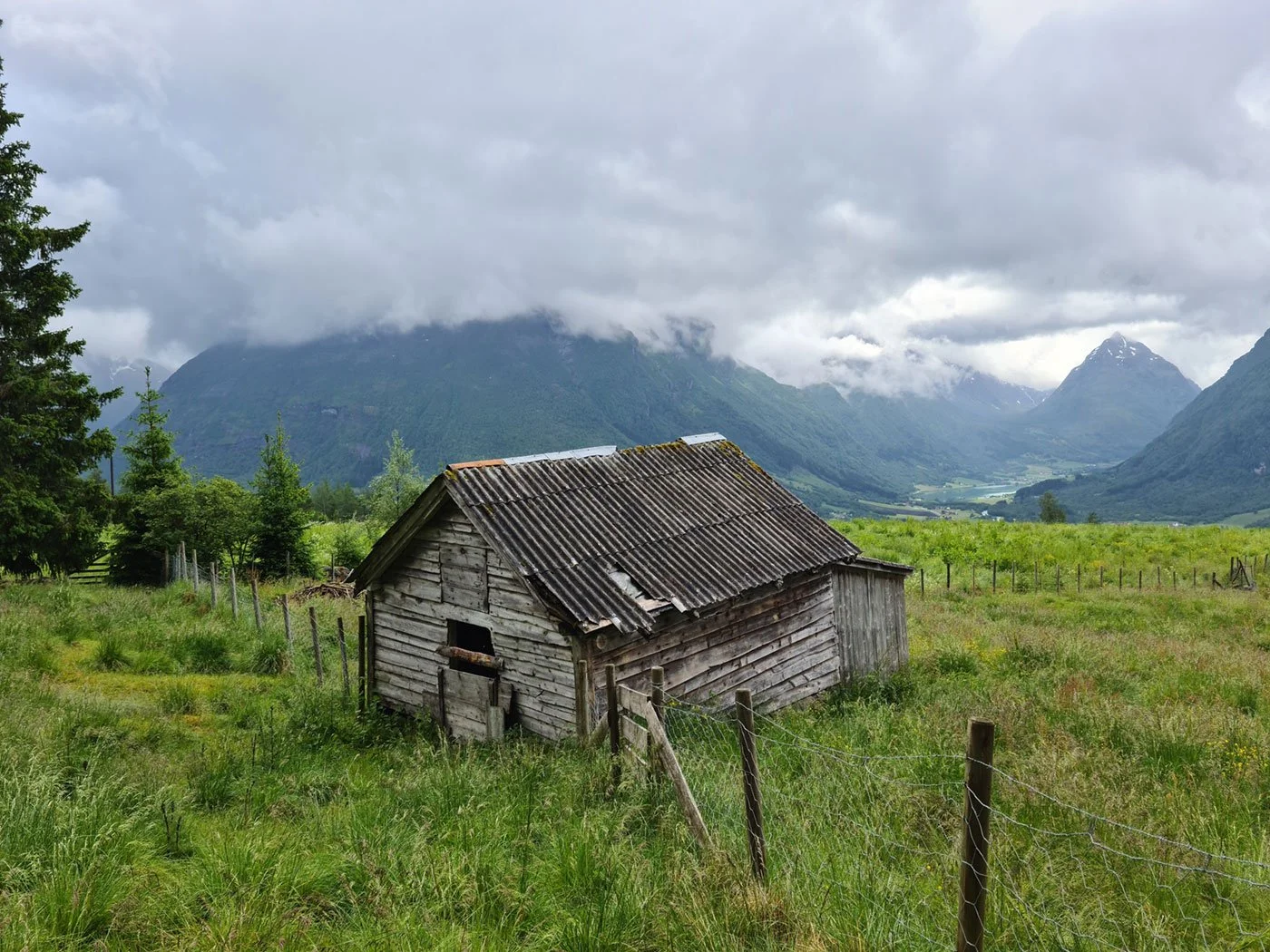 Et gammelt, værslitt vedskal i en grønn eng med fjell og skyer i bakgrunnen.
