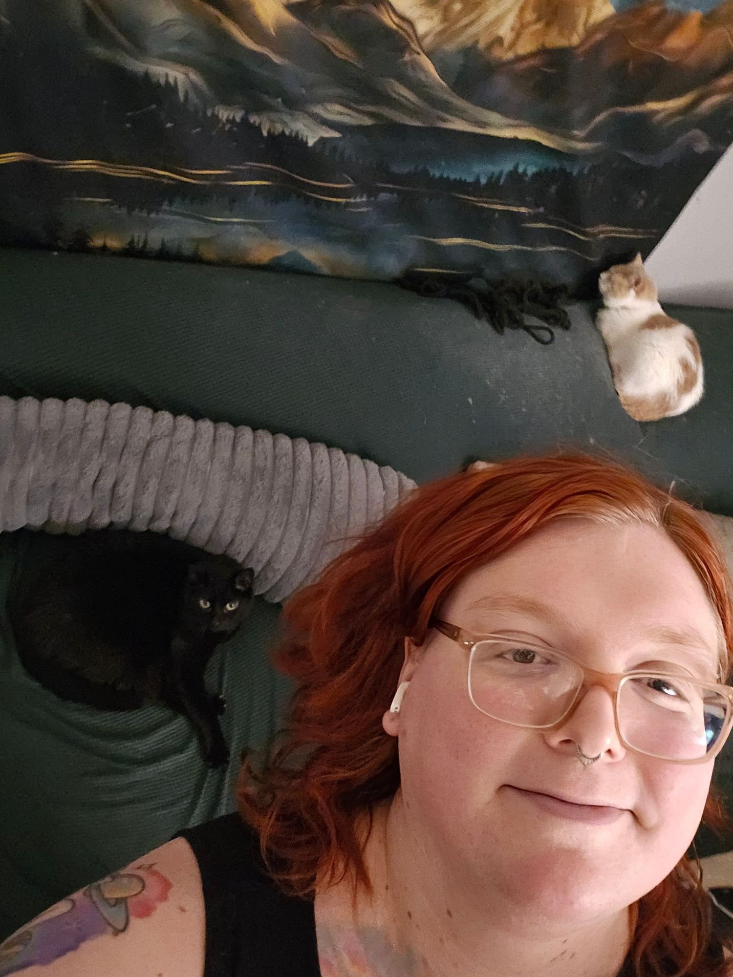 A woman with red hair, glasses, and light skin taking a selfie, with two cats lying on her and a dark couch behind her. The cats are a black one and a calico with orange, white, and black fur.
