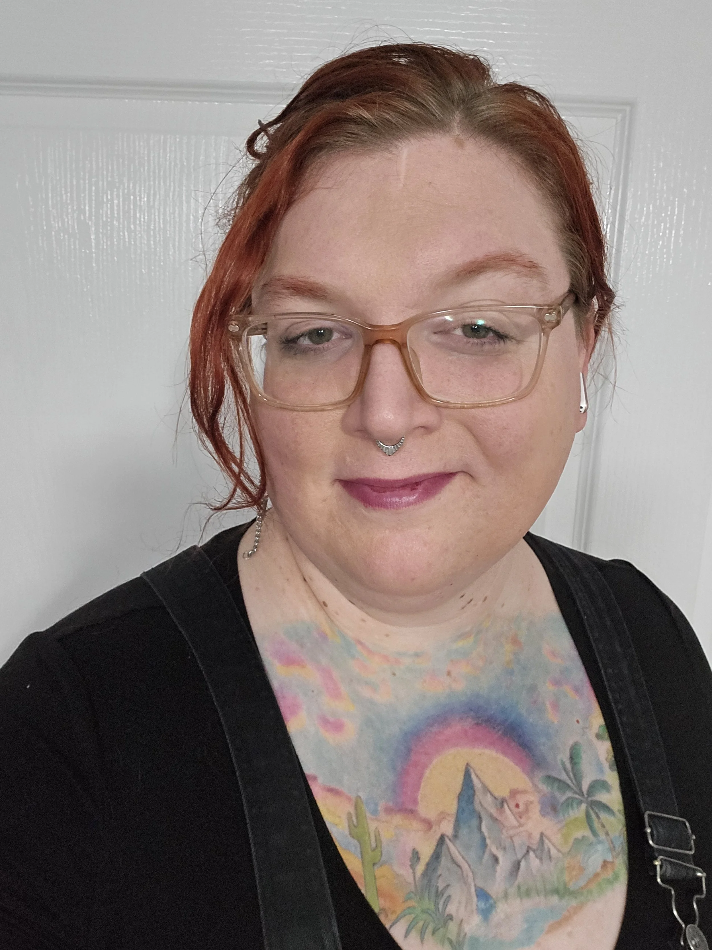 A woman with red hair, wearing glasses and a colorful shirt with a mountain and sunset scene, standing in front of a white textured door.