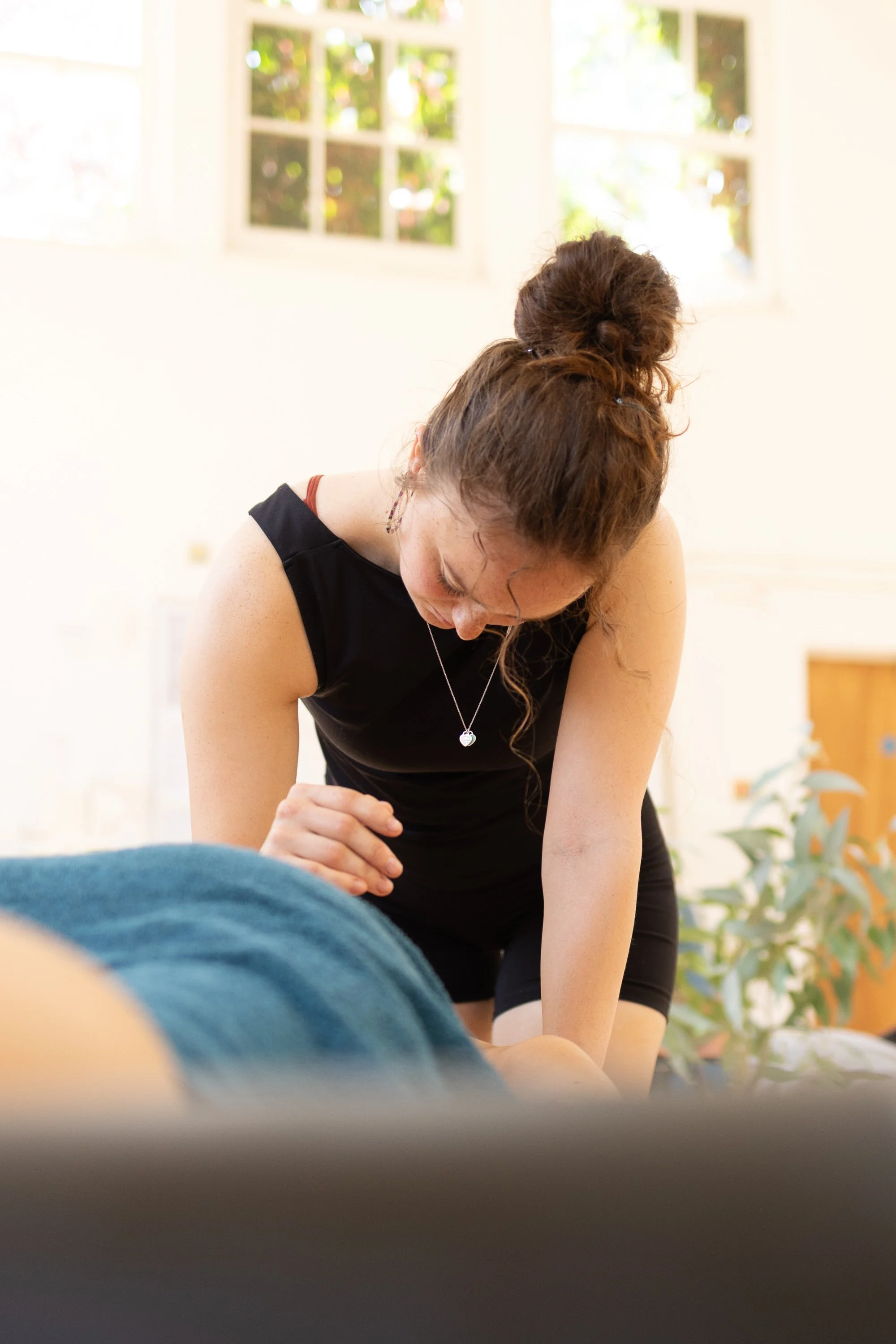 A woman with brown hair tied in a bun is practicing some form of massage or therapy, focusing on a person's leg. The woman is wearing a black sleeveless top and has a silver heart-shaped necklace. The setting appears to be a bright room with large windows and some greenery visible outside.