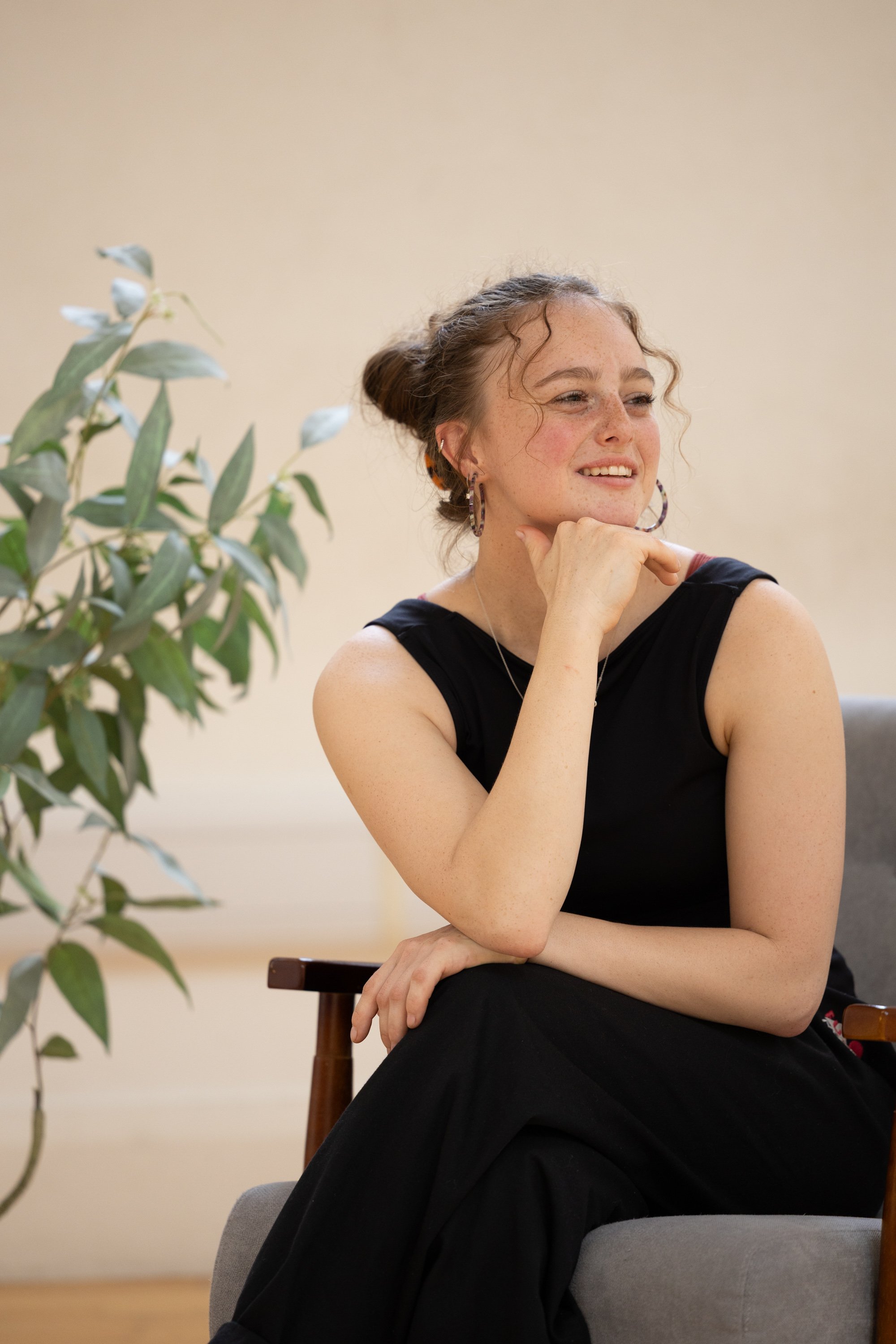 A young woman with curly hair and freckles smiling, sitting with her chin resting on her hand, wearing a black sleeveless top and large hoop earrings, next to a green leafy plant in an indoor setting.