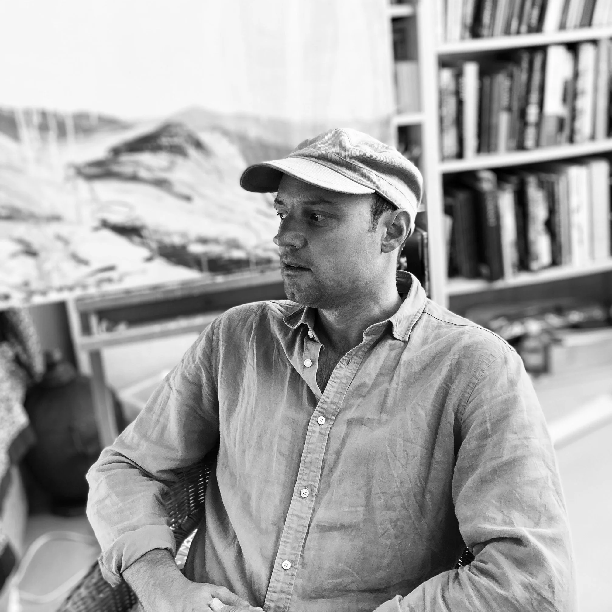 Black-and-white photo of a young man wearing a cap and a button-down shirt, sitting indoors with a bookshelf and artwork in the background.