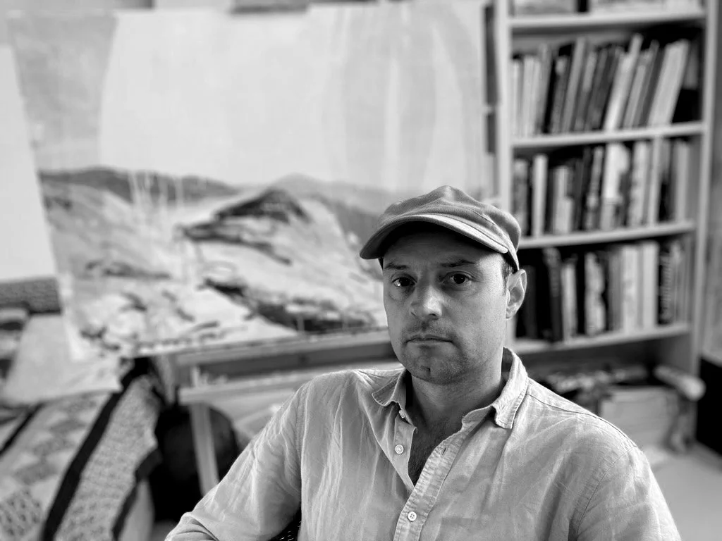 A man wearing a cap and a button-up shirt in a room with bookshelves and a large painting of mountains in the background.