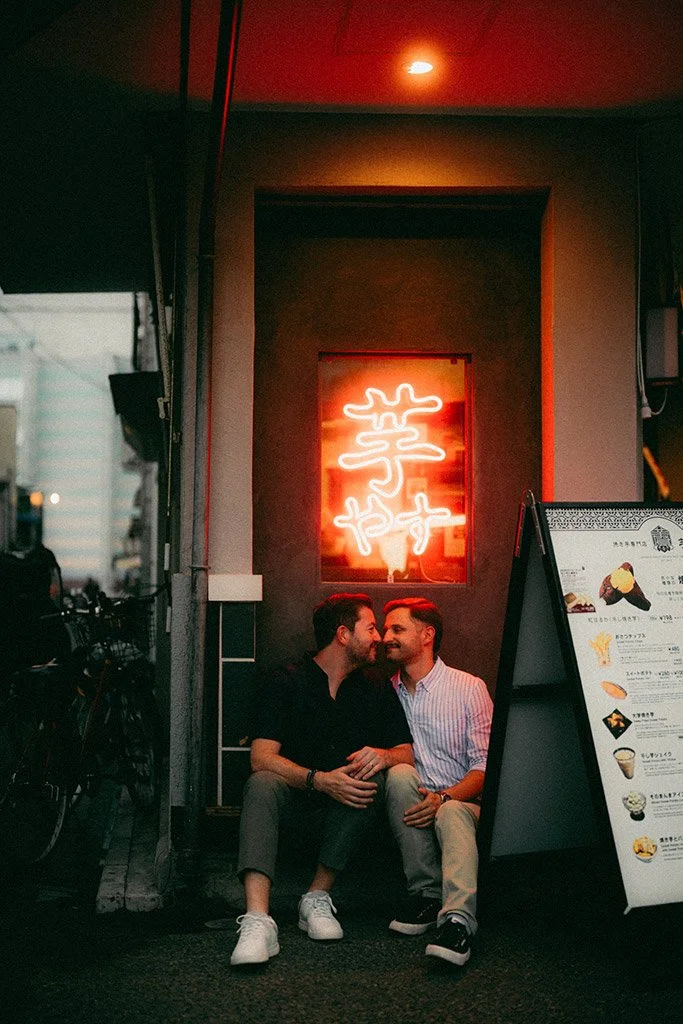 Two men sitting on the sidewalk outside a restaurant with a neon sign in an Asian script, smiling and engaged in conversation. There is a menu board beside them and bicycles parked nearby.