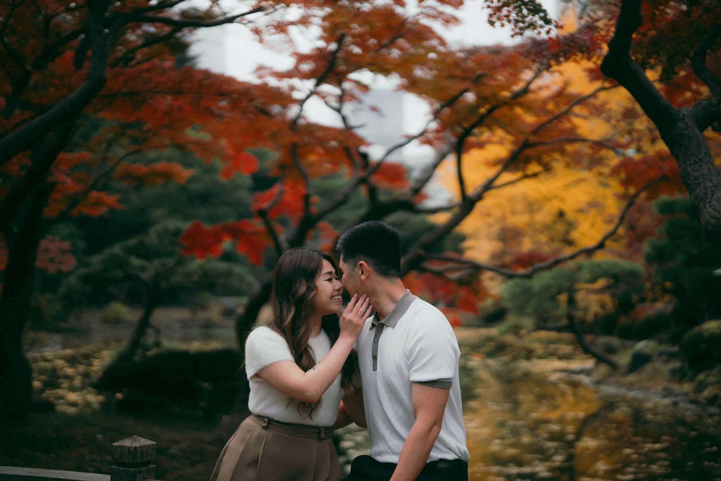 A couple stands close together outdoors amid autumn trees with vibrant red, orange, and yellow leaves, smiling and looking into each other's eyes.
