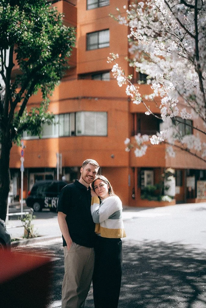 A happy couple standing on a city street with an orange building and blooming cherry blossom trees in the background.