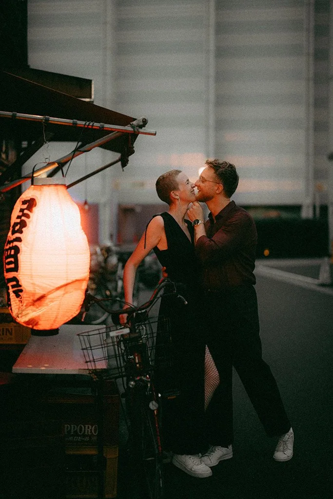 A couple sharing a kiss at night near a food cart with a red lantern, in an urban setting.
